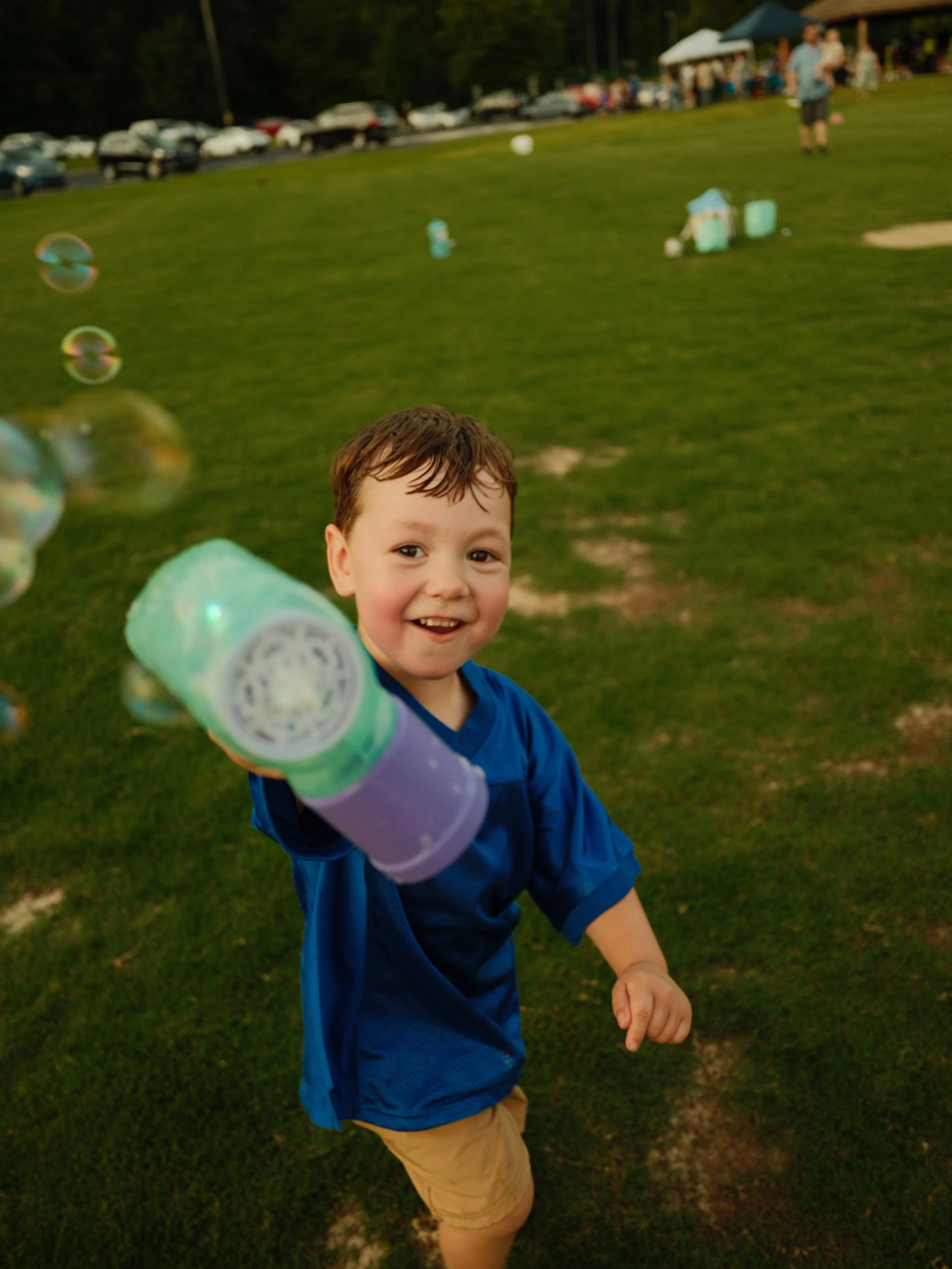 A young boy smiling and holding a bubble-blowing toy towards the camera on a grassy field, with other children and spectators in the background during an outdoor event in the evening.
