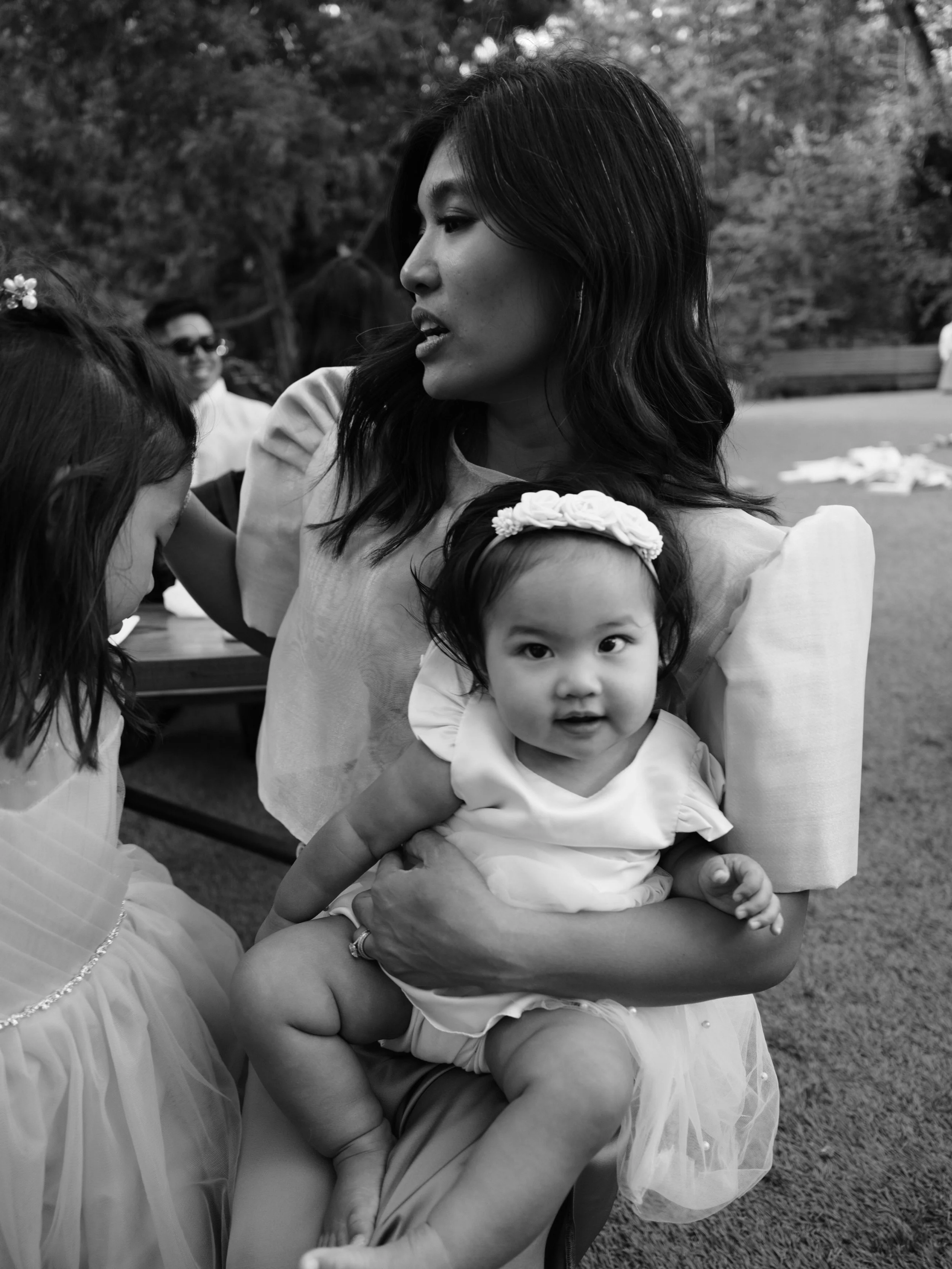 A woman holding a young girl with a floral headband outdoors during the daytime, with other people and trees in the background.