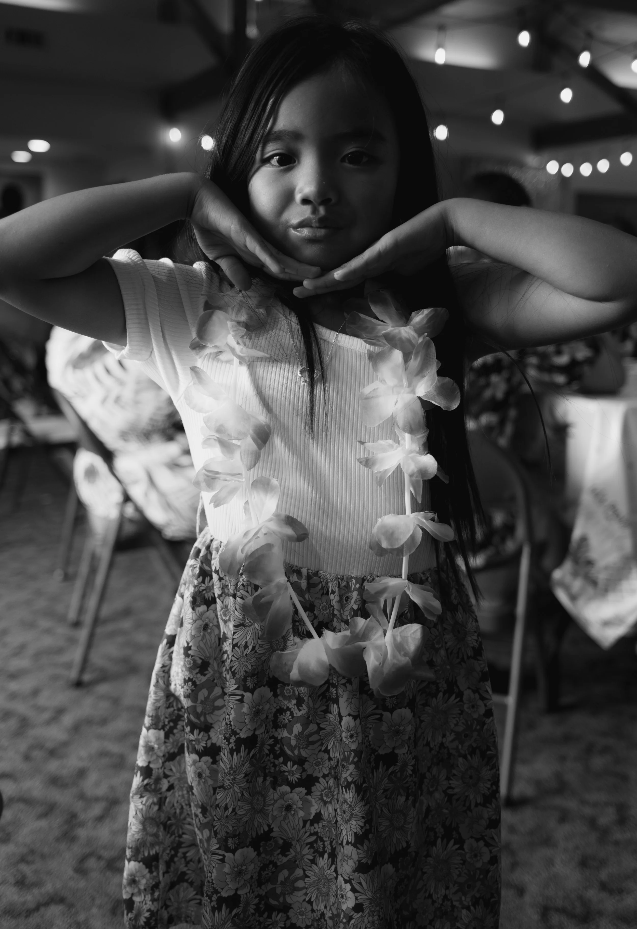A young girl posing with her hands under her chin, wearing a floral lei and a flower-patterned skirt at a festive event with string lights in the background.