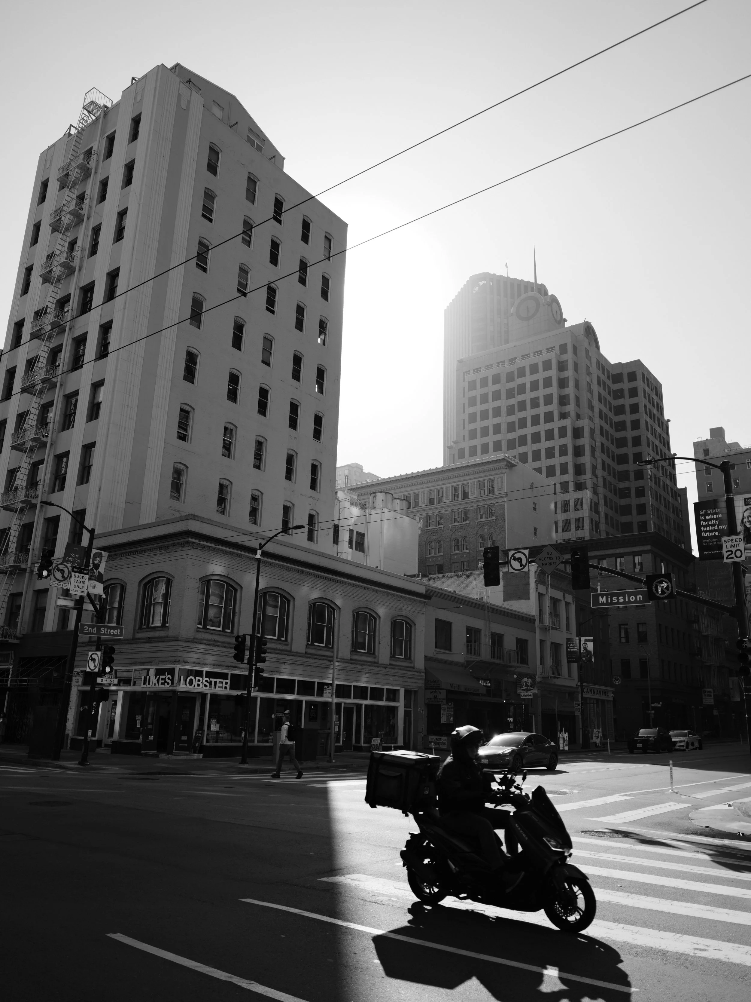 Black and white city street scene with tall buildings, pedestrians, cars, and a person riding a scooter in the foreground.