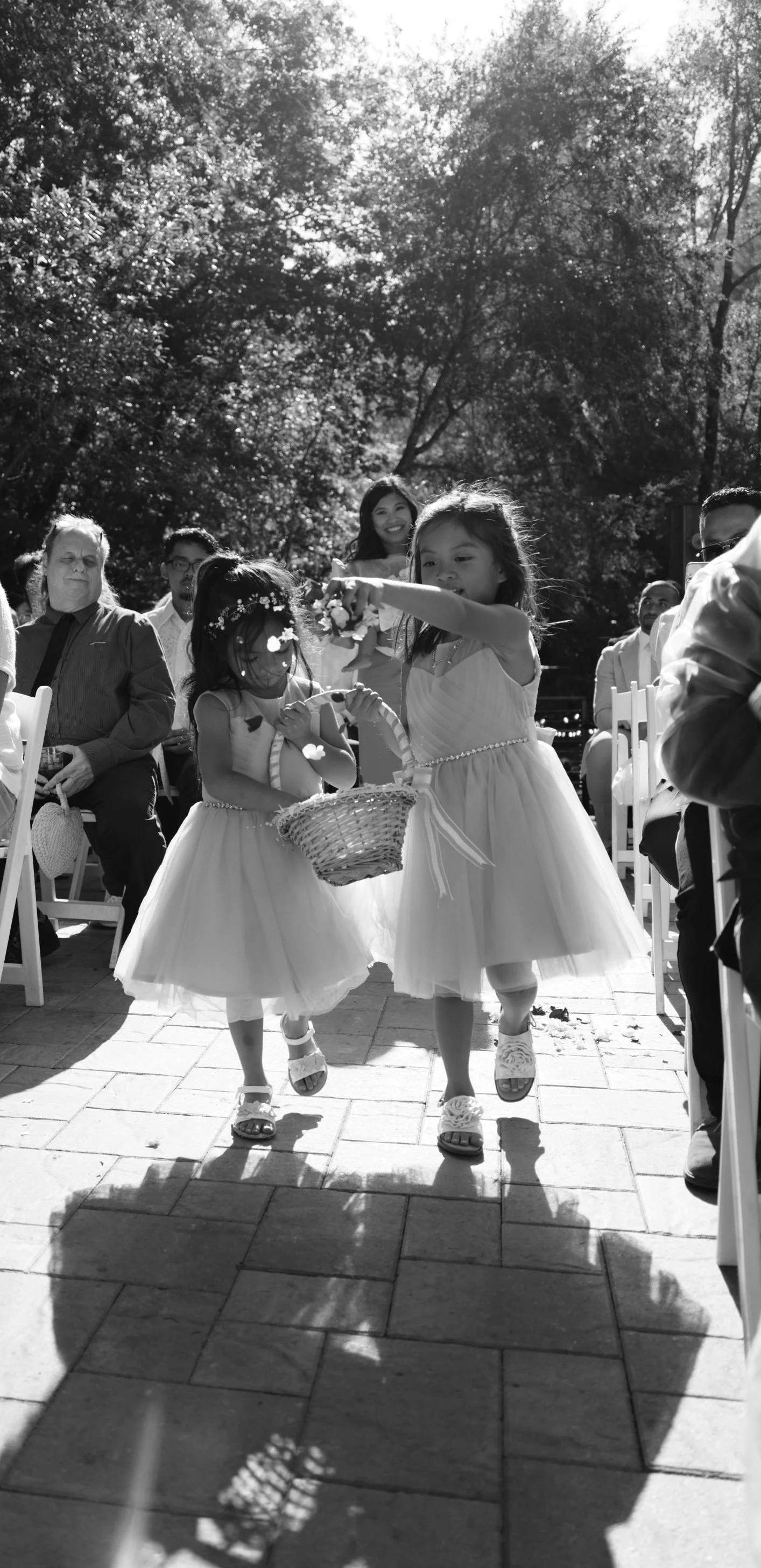 Two young girls in white dresses and sandals walking down an outdoor aisle holding a basket, possibly during a wedding ceremony; adults seated on either side watching, trees and sunlight in the background.