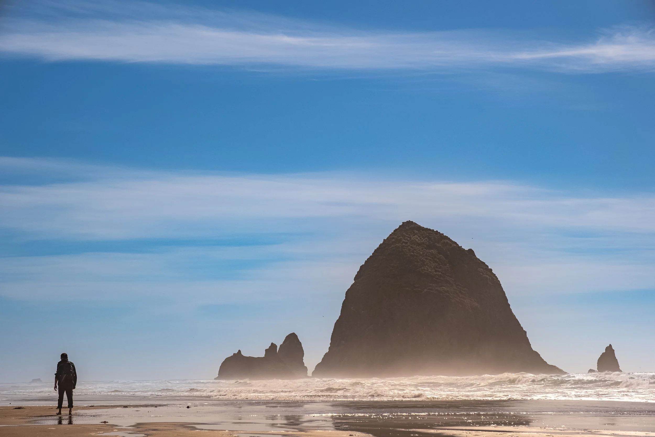 A person standing on a sandy beach facing a large sea stack formation in the ocean, with blue sky and clouds above.