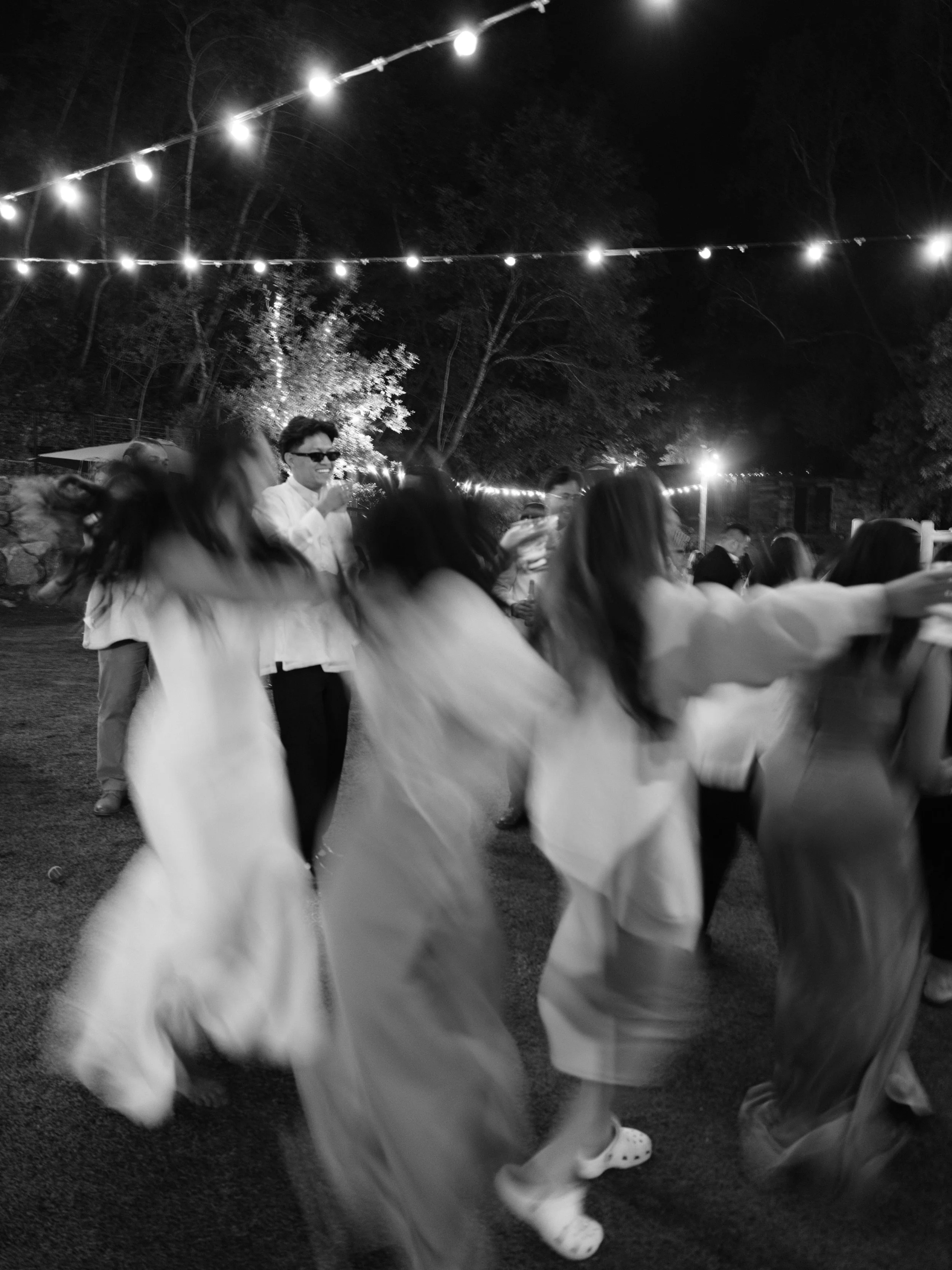 People dancing outdoors at a nighttime celebration under string lights, with some wearing casual clothes and others in dresses.