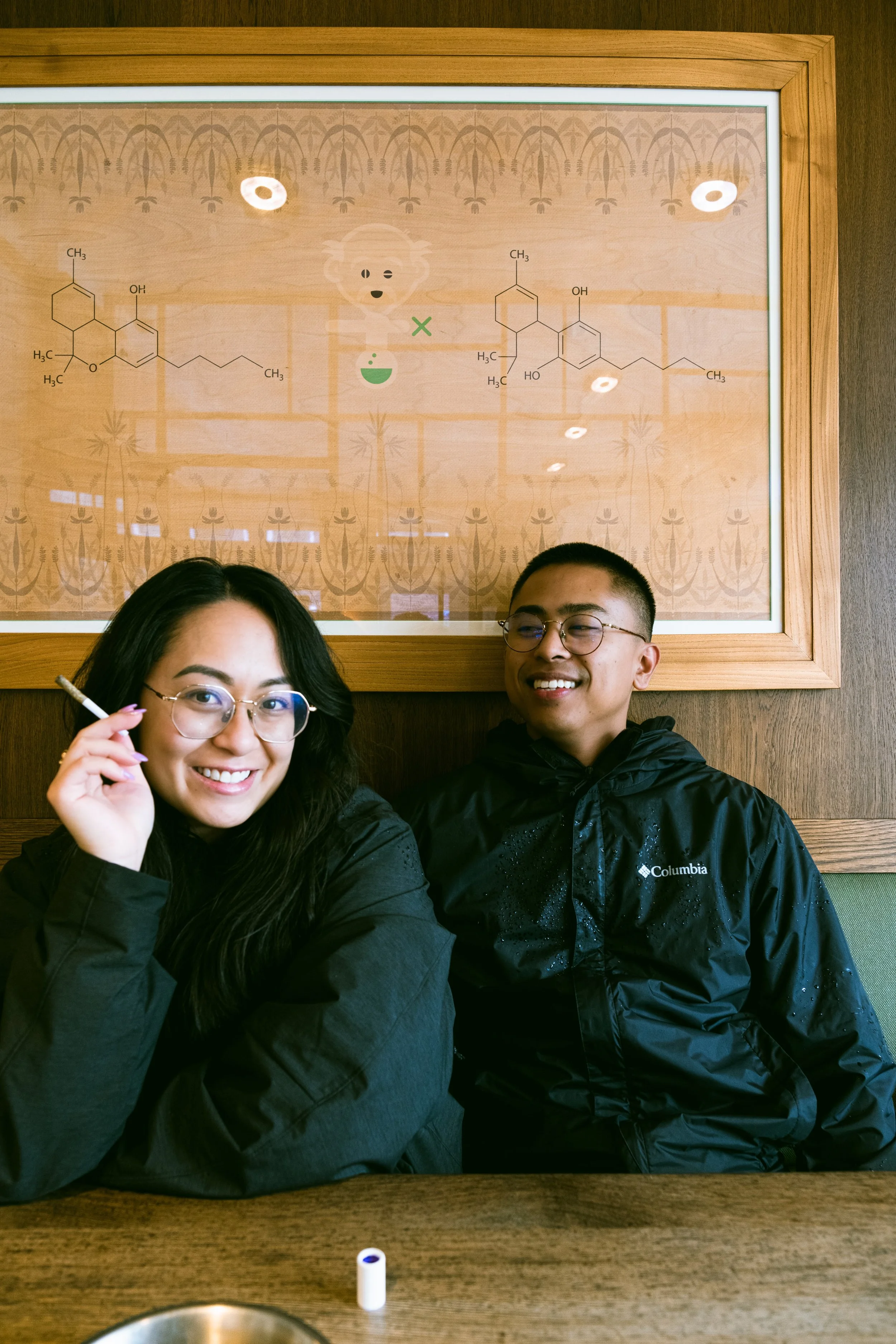 Two young people sitting at a table in a restaurant, smiling, with a molecular structure diagram on a framed board behind them.