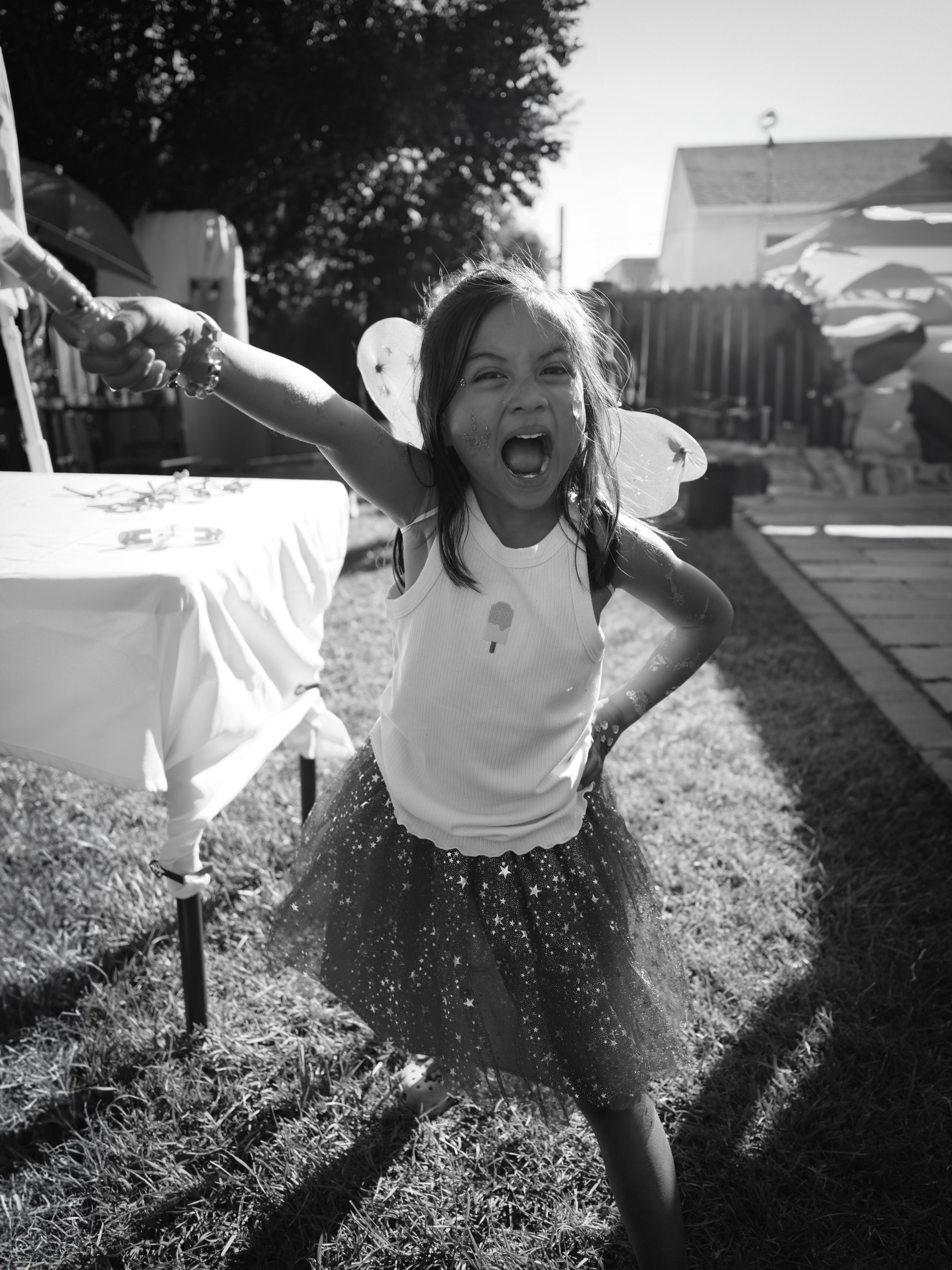 A young girl dressed as a fairy with butterfly wings, wearing a starry skirt, excitedly reaching out with her arm during a birthday party outside.