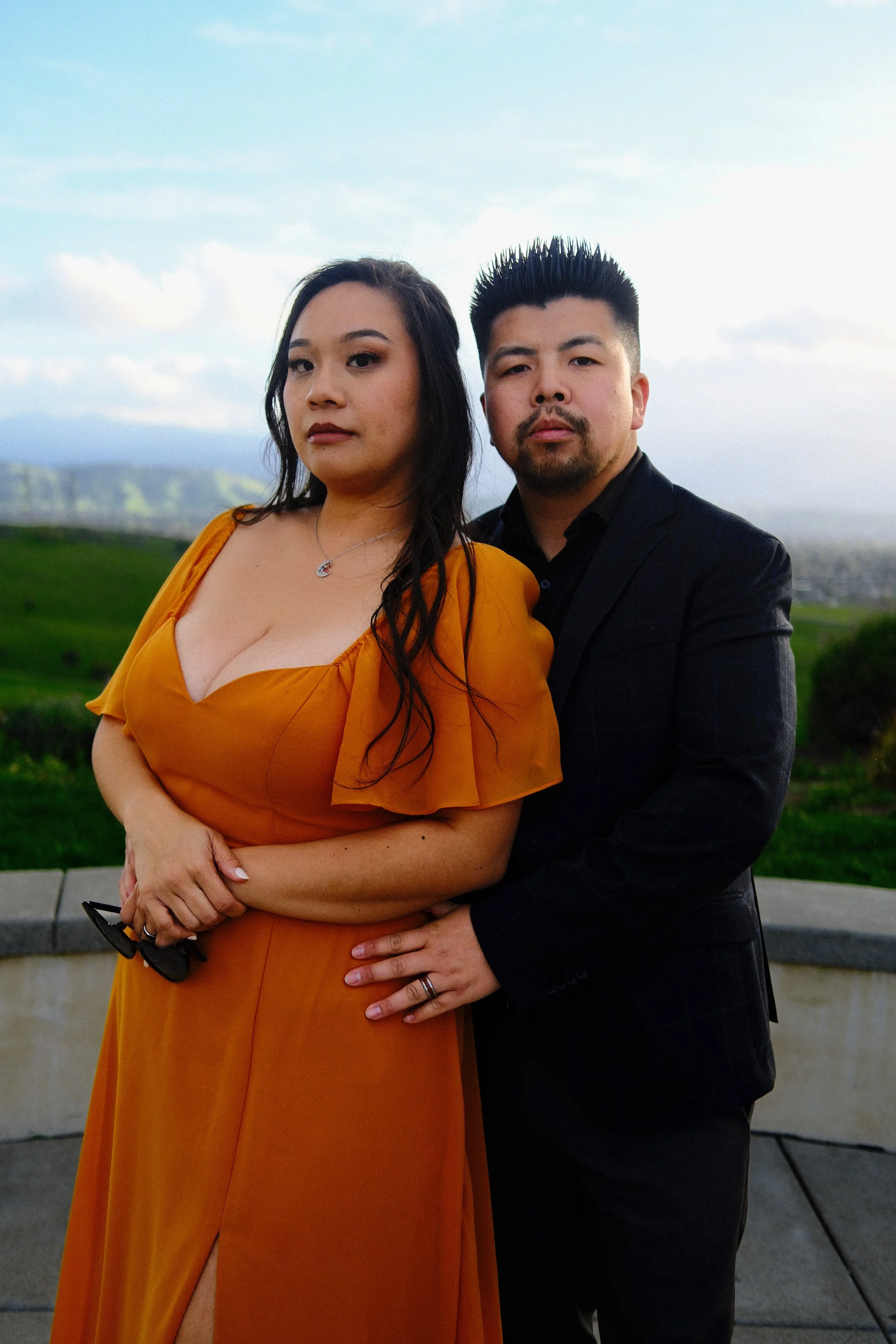 A man and woman standing outdoors with greenery and hills in the background, both looking at the camera. The woman is wearing an orange dress with a slit, and the man is dressed in a dark suit.