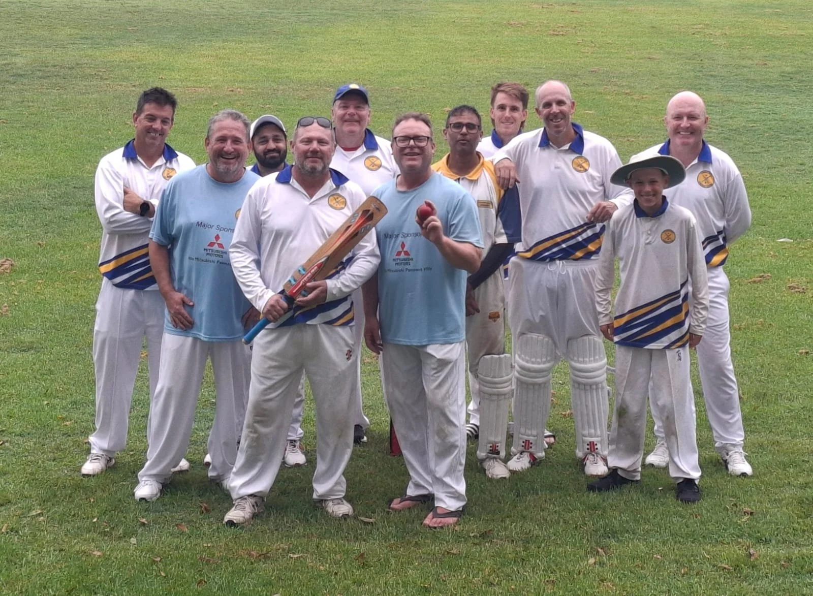 A group of cricket players in white and blue uniforms posing on a grassy field, some holding cricket equipment, smiling at the camera.
