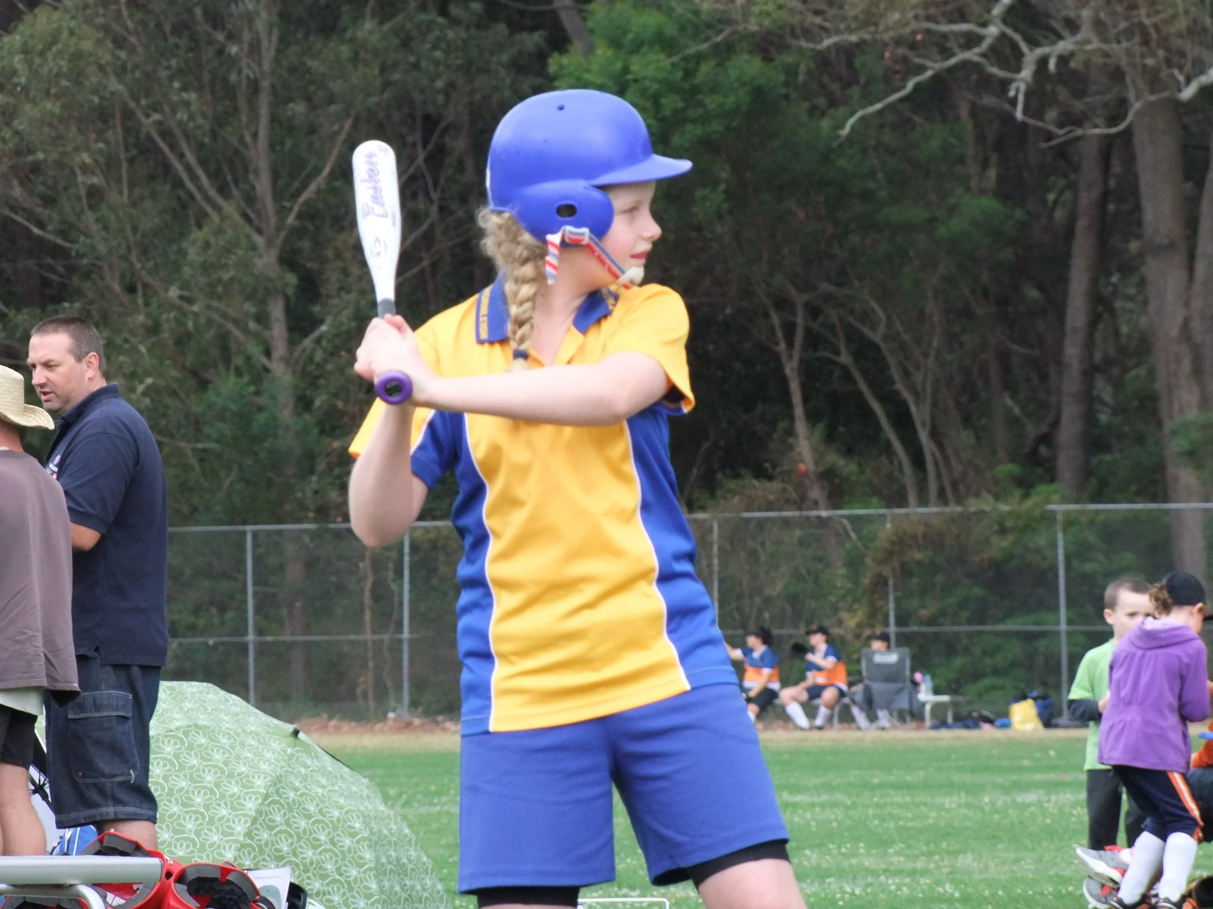 A young girl dressed in yellow and blue sports uniform, wearing a helmet, holding a baseball bat, preparing to swing during a game on a baseball field.