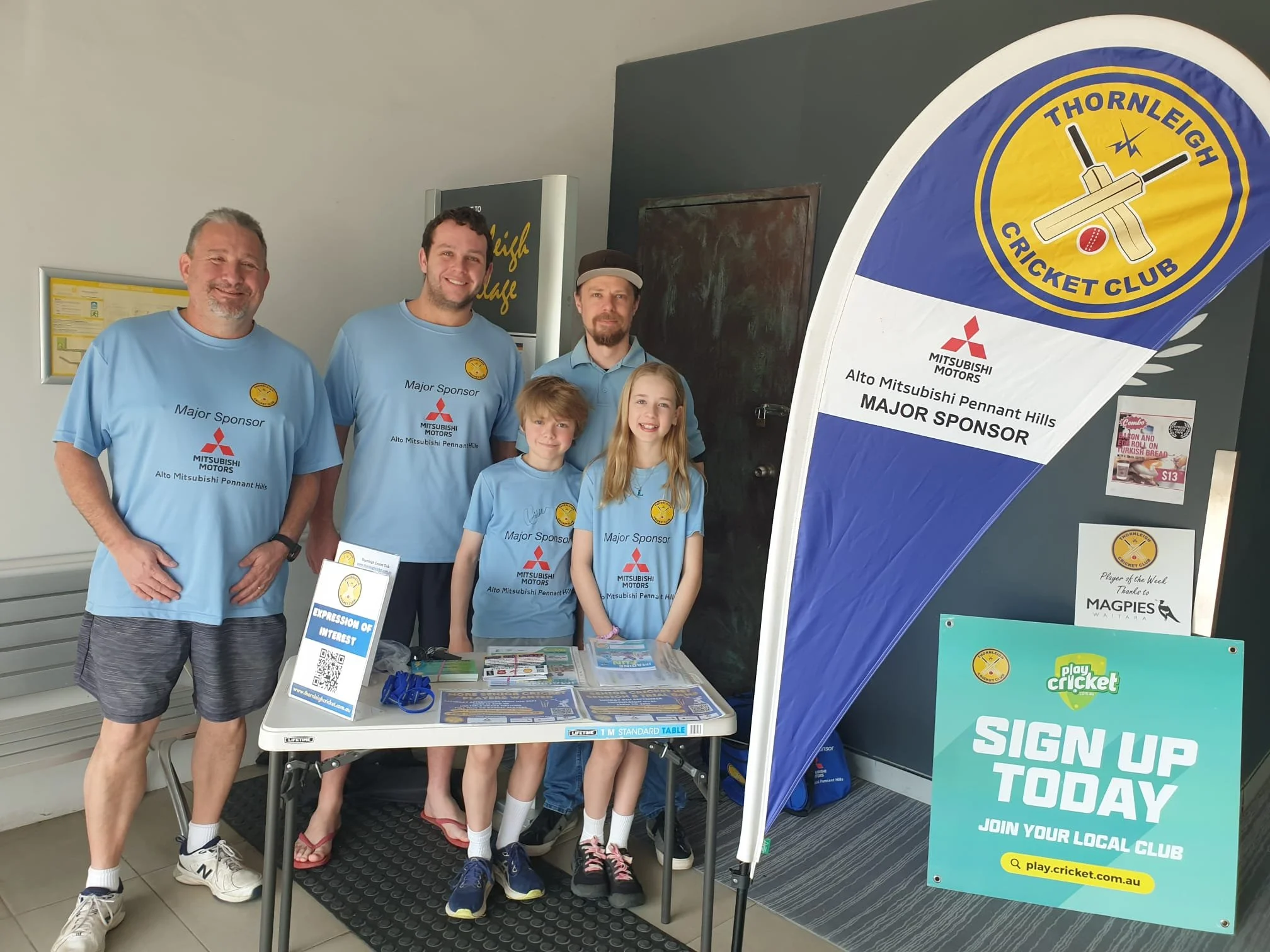 A group of five people standing behind a table at a cricket club booth, wearing blue shirts with sponsorship logos, with a large promotional banner. The booth promotes signing up for a local cricket club.