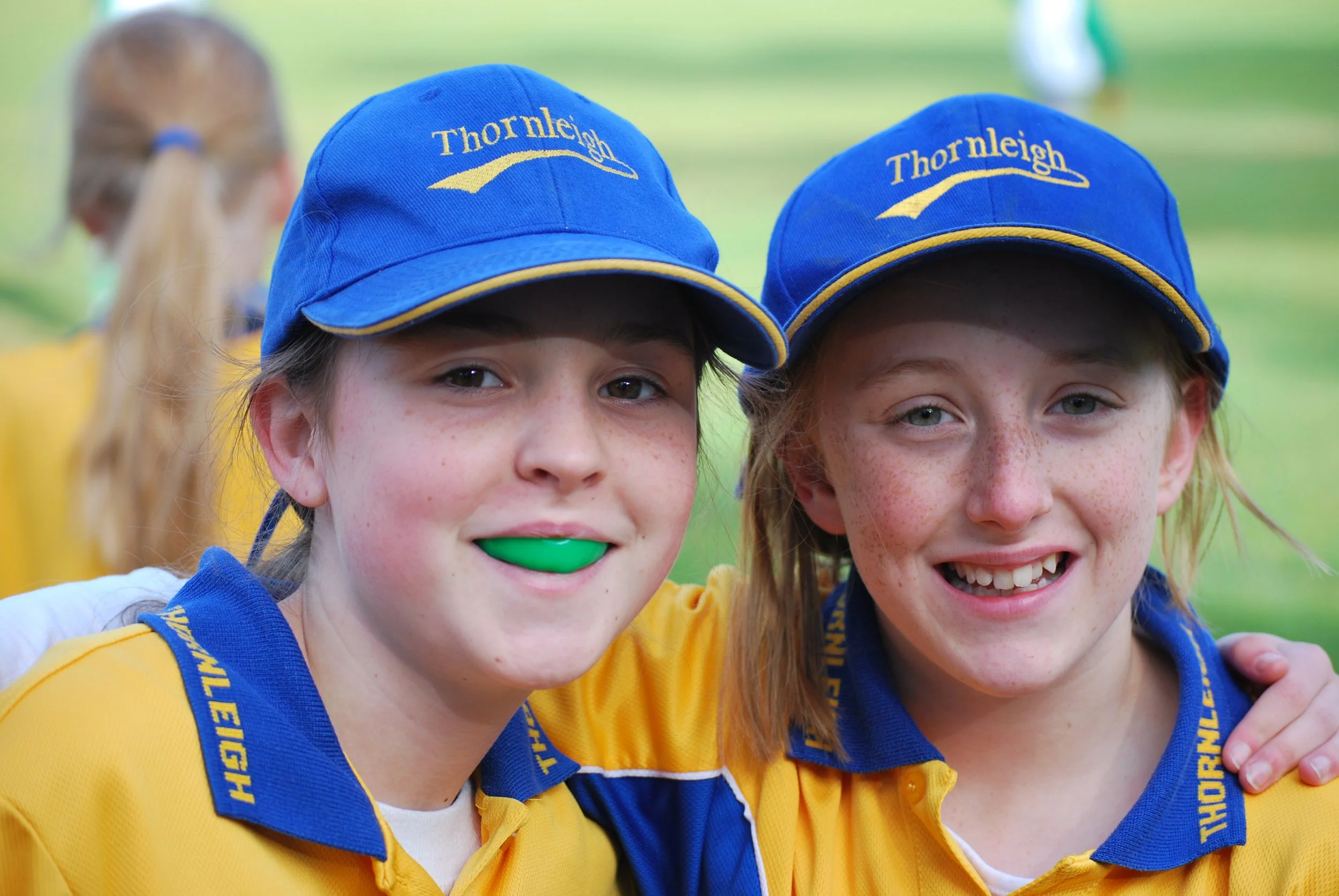 Two young girls wearing blue caps with 'Thornleigh' embroidered on them and yellow sports uniforms, smiling and posing together outdoors.