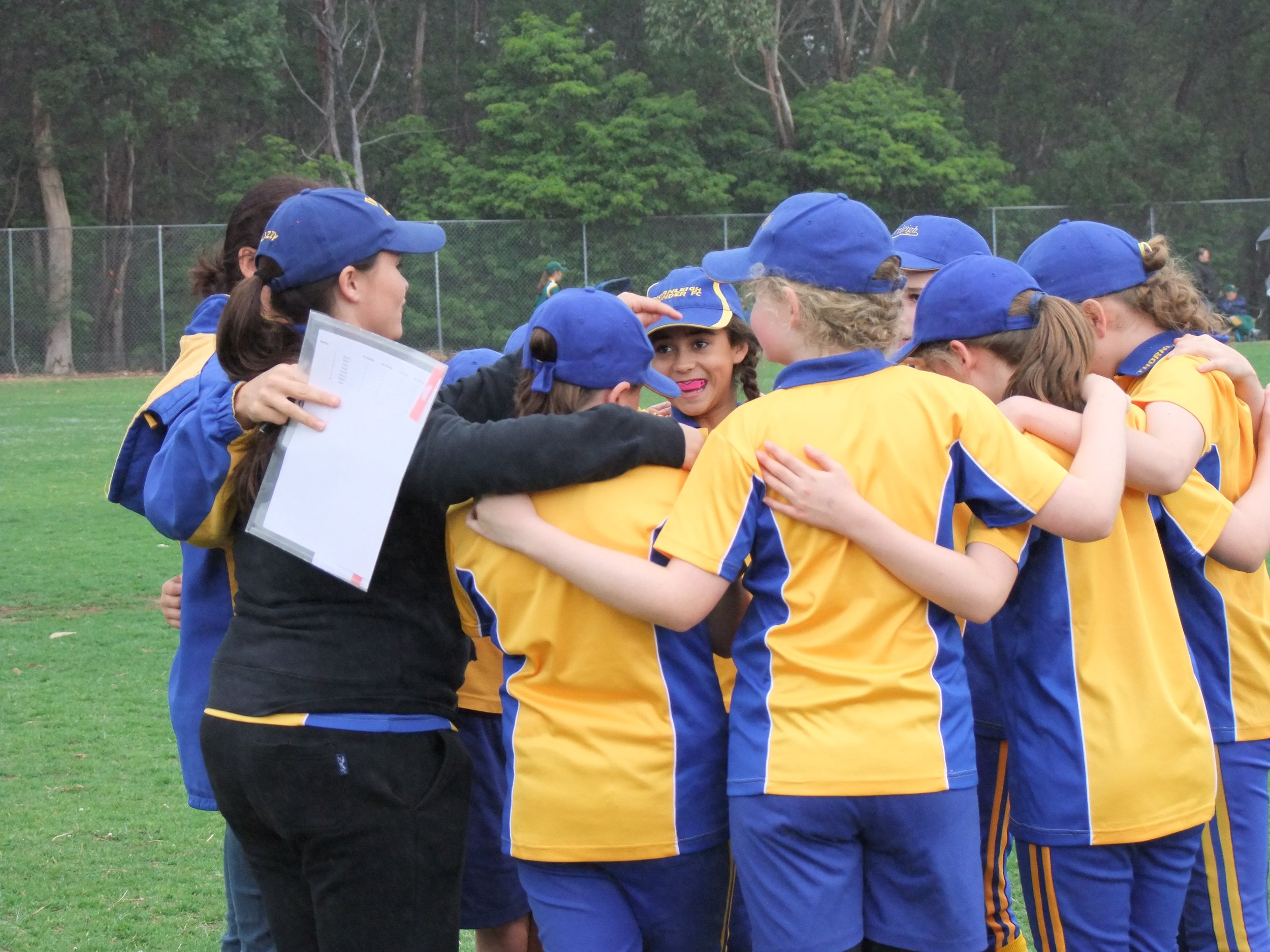 A group of young female soccer players, dressed in matching yellow and blue uniforms and hats, are gathered in a huddle on a soccer field, with their coach holding a clipboard nearby.