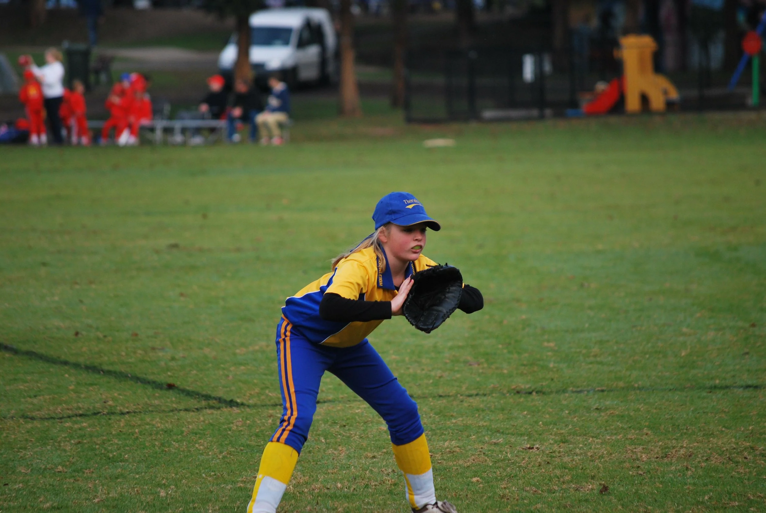 A young girl in a yellow and blue sports uniform, wearing a blue cap and yellow socks, playing softball on a grassy field.