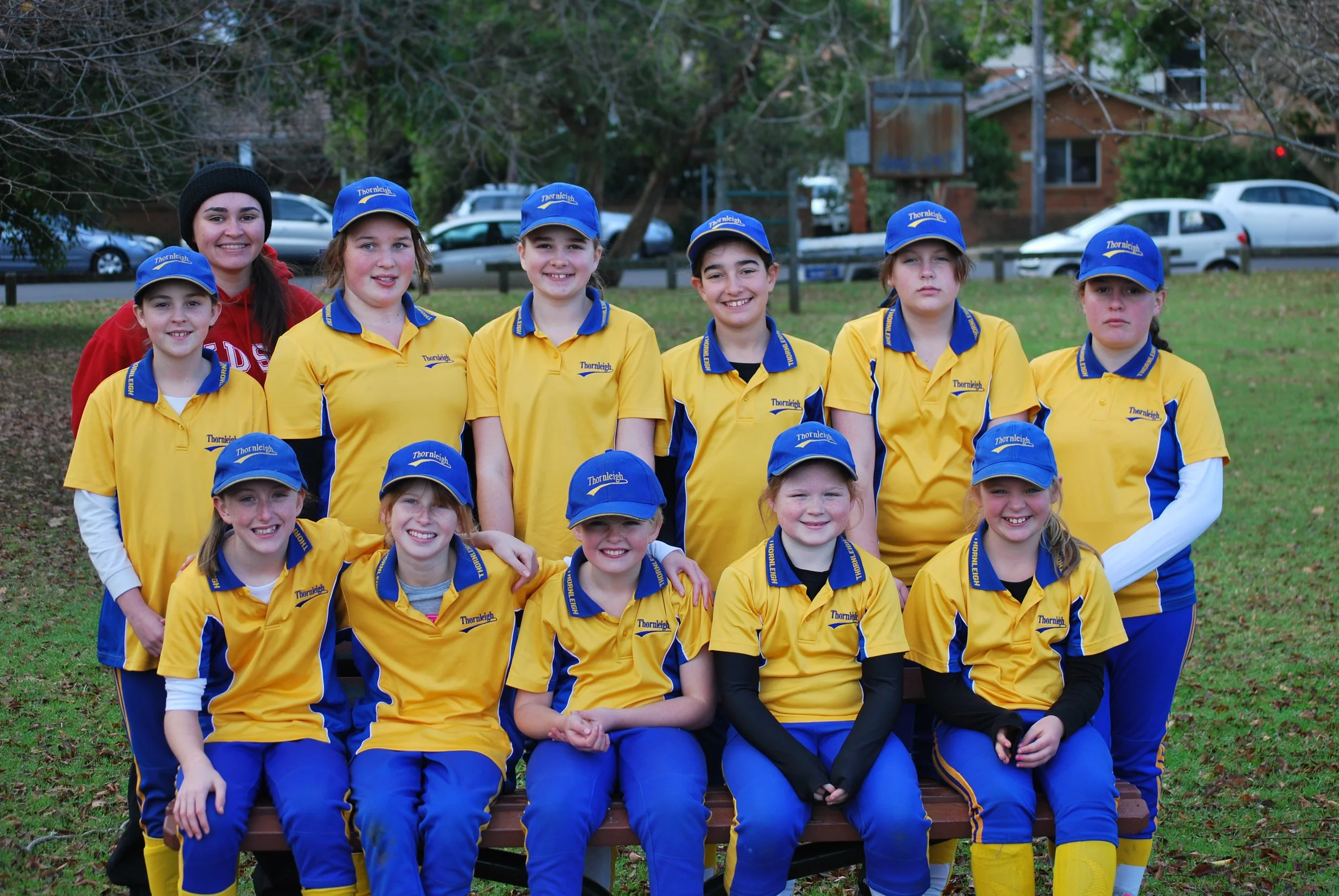 A group of young female softball players in yellow and blue uniforms with a coach or adult in red attire, posing outdoors on a bench in a park.