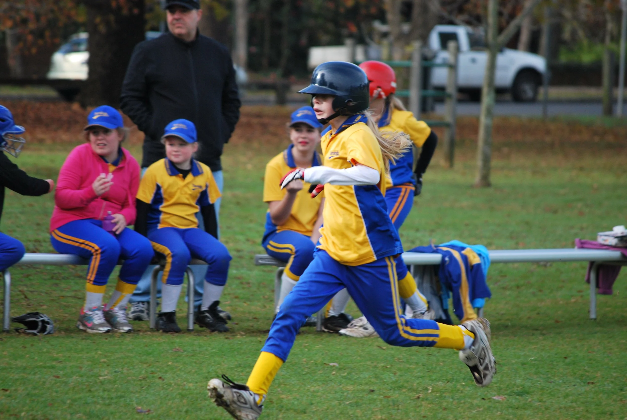 Young girl in yellow and blue sports uniform running on a field during a youth sports game, with other children and a coach watching from the sideline.