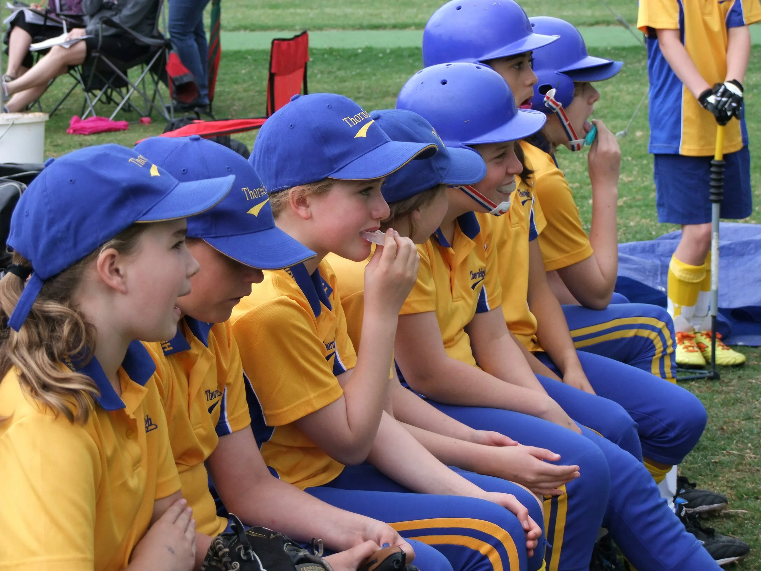 Youth baseball team wearing yellow and blue uniforms sitting on a bench, some wearing blue helmets, watching a game.