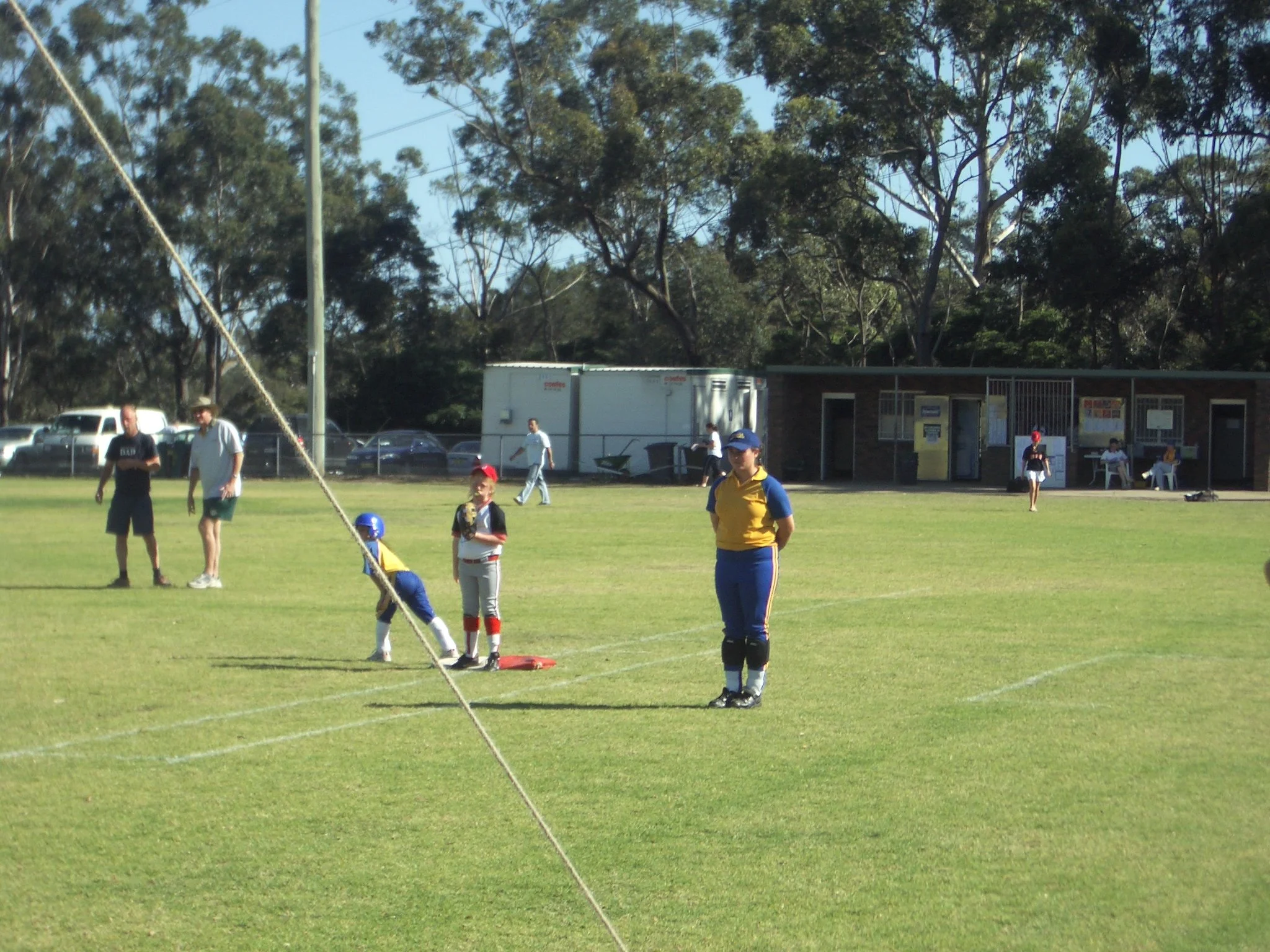 Children and coaches on a grassy field during a baseball practice or game with a small brick building and trees in the background.