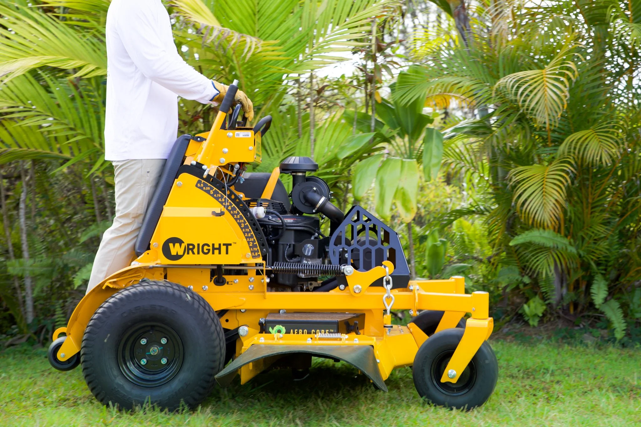 A person operating a yellow Wright aerator machine on a grassy area with green foliage in the background.