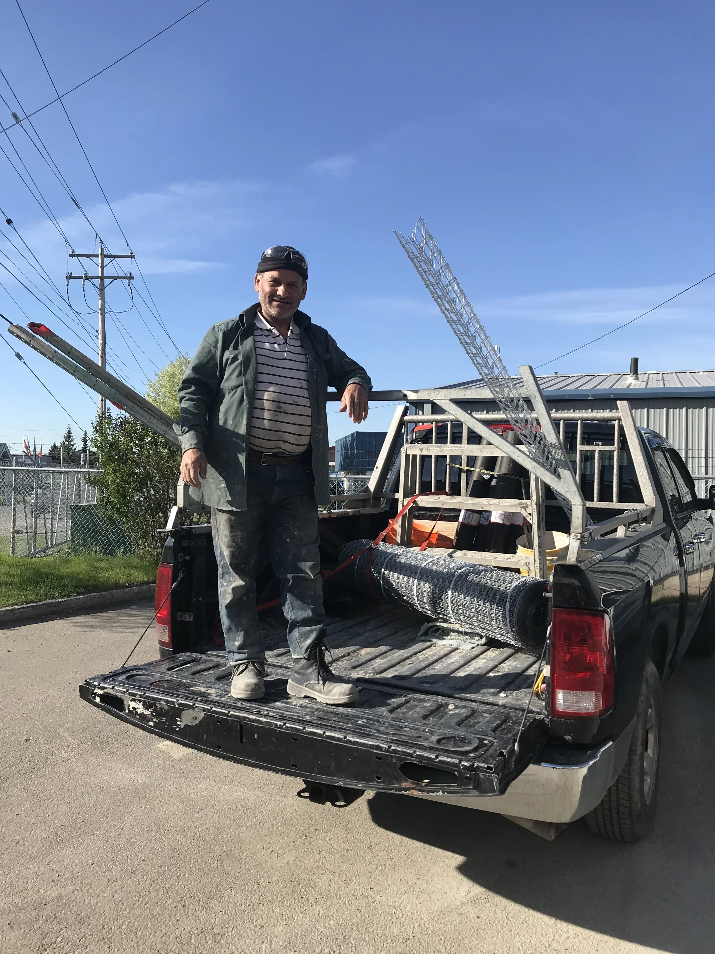 A man standing in the back of a pickup truck with equipment, wearing work clothes and a cap, outdoors on a sunny day. There are power lines and a building in the background.