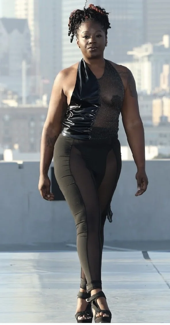 A woman walking on a rooftop with city buildings in the background, wearing a black halter top with leather and sparkly details, black sheer leggings, and black high-heeled sandals.