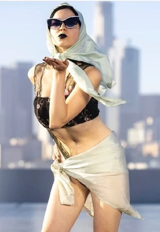 A woman in a vintage-style scarf, dark sunglasses, and dark lipstick standing outdoors with a city skyline in the background.