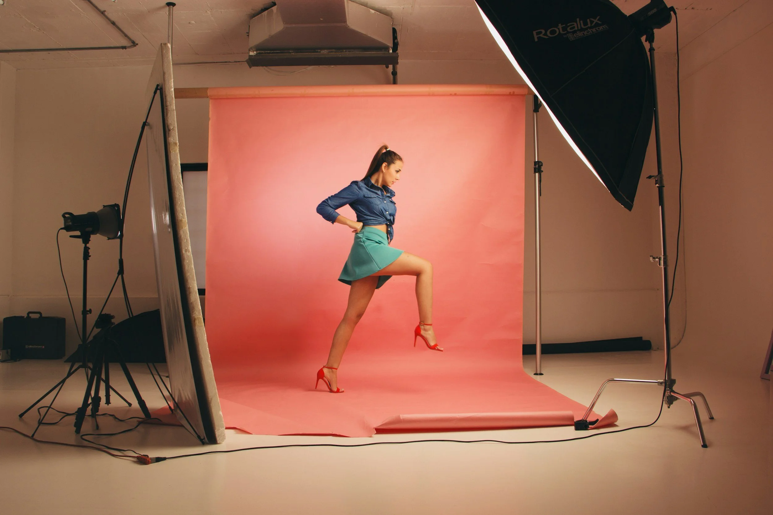 A woman posing in a photography studio with a pink backdrop, wearing a blue shirt, teal skirt, and red high heels.