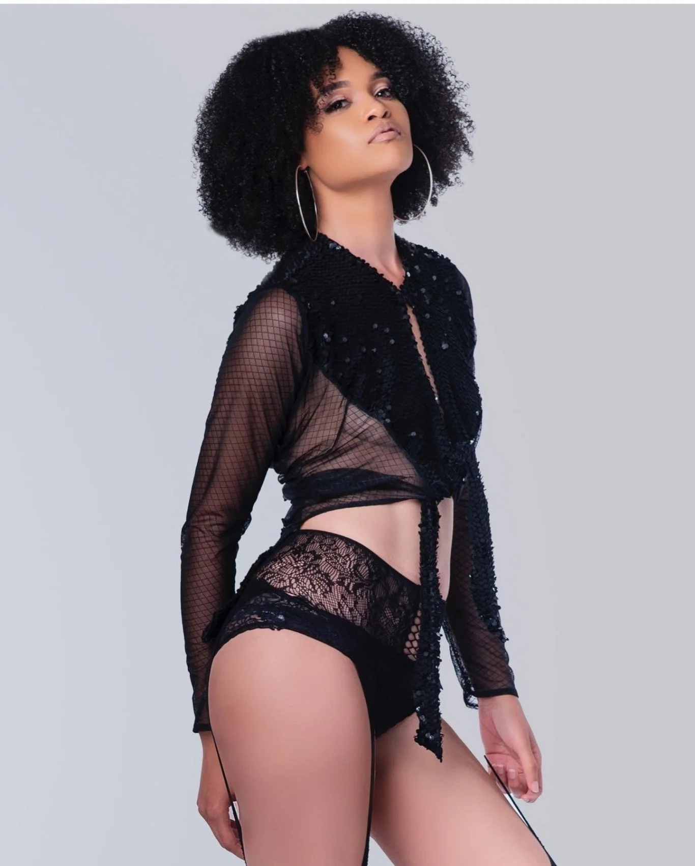 A woman with curly black hair wearing black lace and mesh lingerie, standing against a plain light gray background.