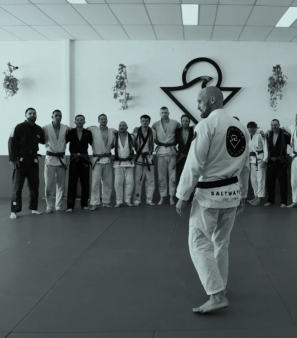 A group of Brazilian Jiu-Jitsu practitioners standing in a line with their instructor in the foreground. The practitioners are wearing gi uniforms with various belt colors. The instructor is facing the group and is dressed in a gi with the word 'Saltwater' on the back. The background features a logo on the wall and some decorative plants.