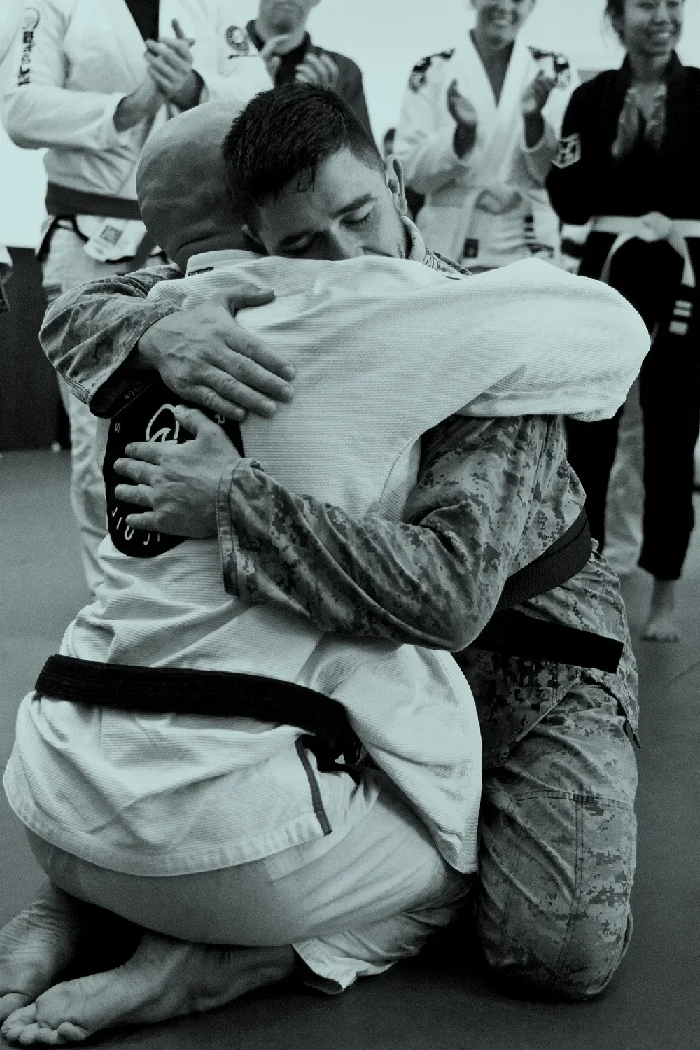 Two martial artists hugging during or after a Brazilian Jiu-Jitsu competition, with others clapping and smiling in the background.