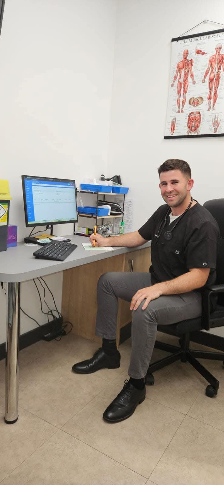 A smiling healthcare professional wearing black scrubs and black shoes sitting at a desk in an medical office. There is a computer monitor, a keyboard, and a shelf with medical supplies on the desk. A muscular anatomy chart is hanging on the white wall behind him.