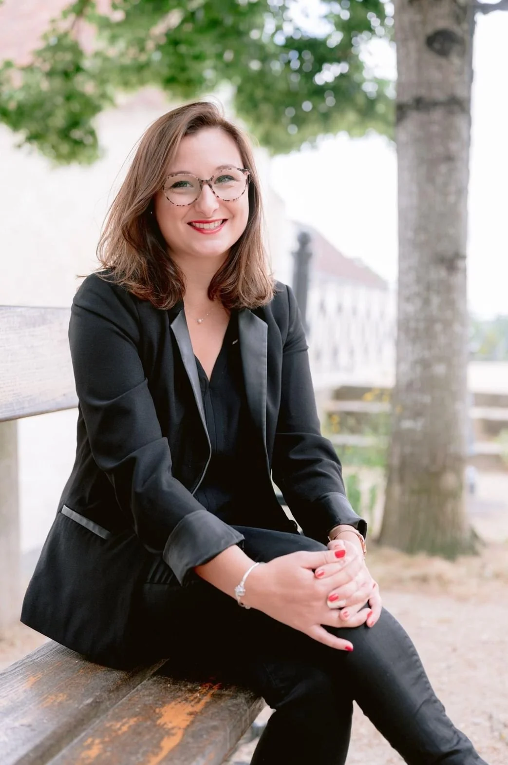 Une femme souriante avec des lunettes, portant un blazer noir, assise sur un banc en bois en plein air avec des arbres en arrière-plan.