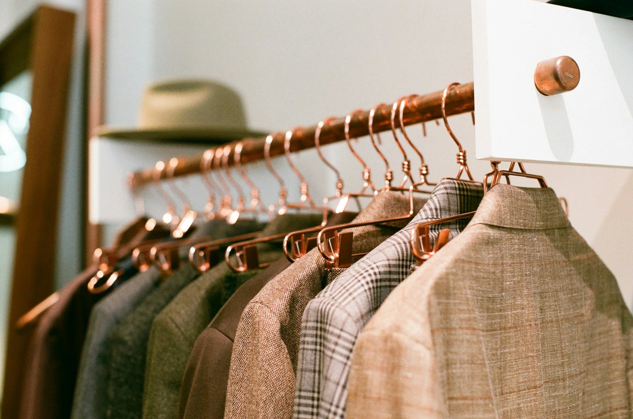 A row of men's blazers hanging on a copper clothing rack, with a beige hat on a shelf above.
