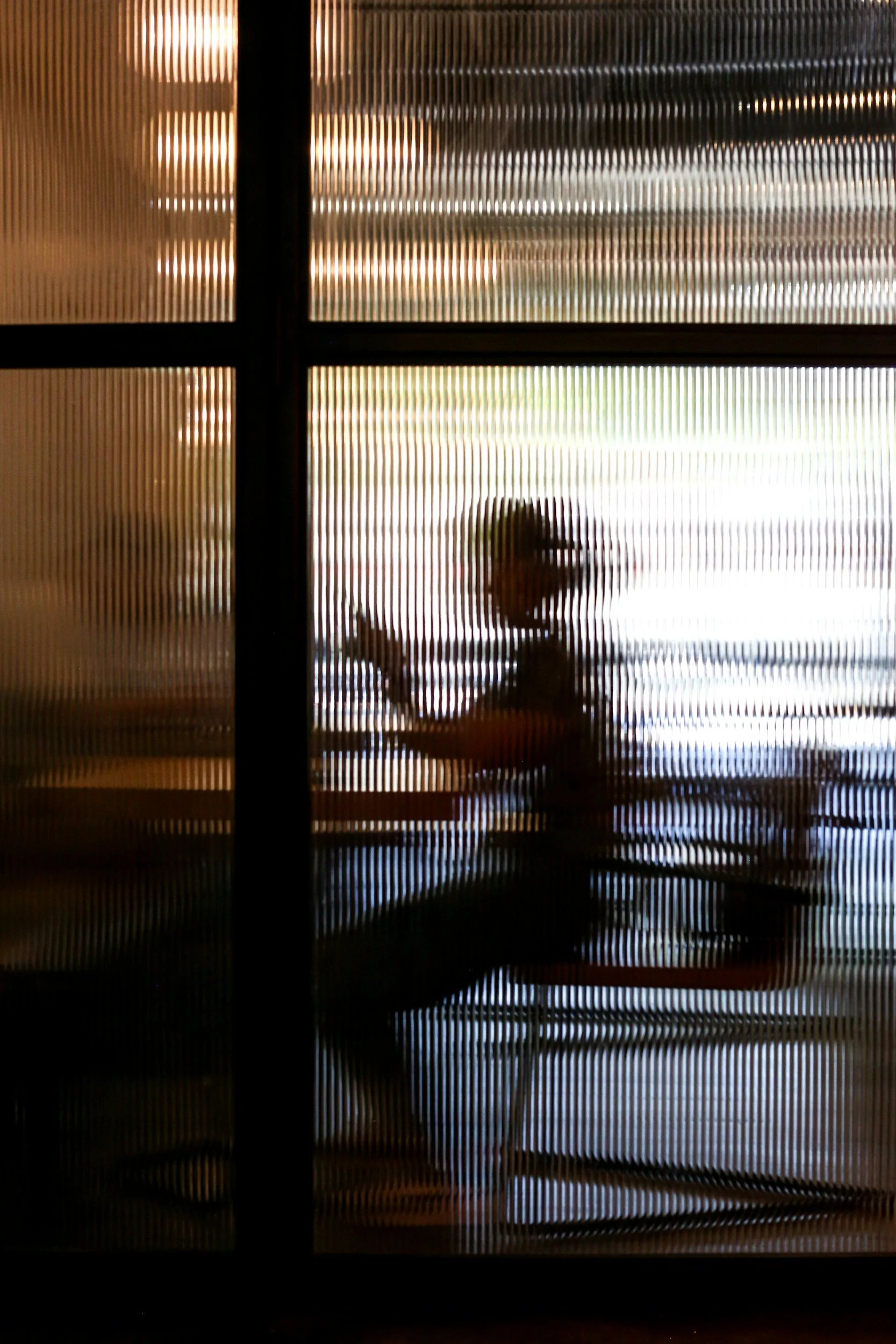 A person sitting at a table, seen through a textured glass or window with vertical lines, making the person appear blurred and obscured.