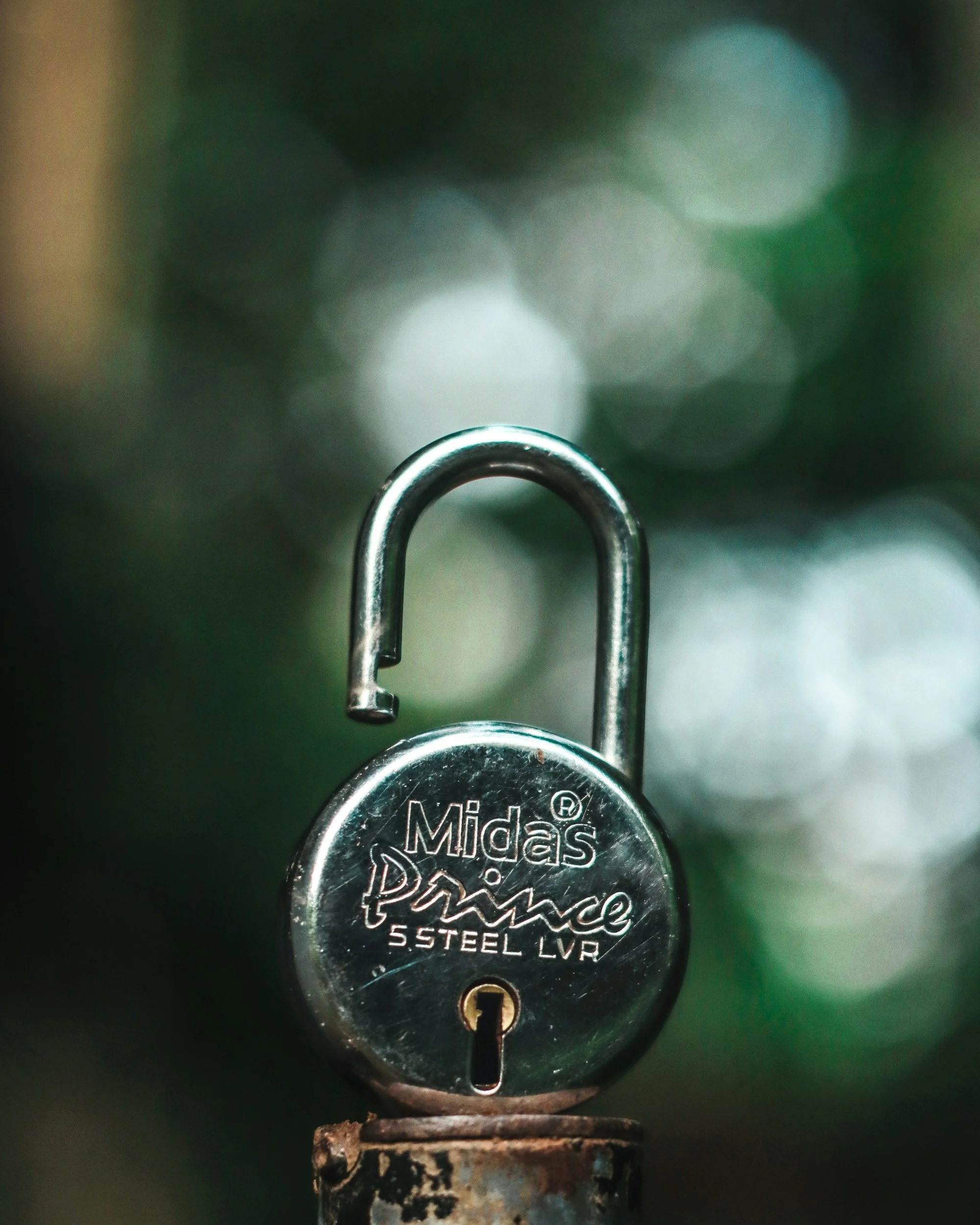 Close-up of a silver padlock with the brand name 'Midas Prime' and '5 Steel LVR' engraved on it, hanging on a rusty surface.