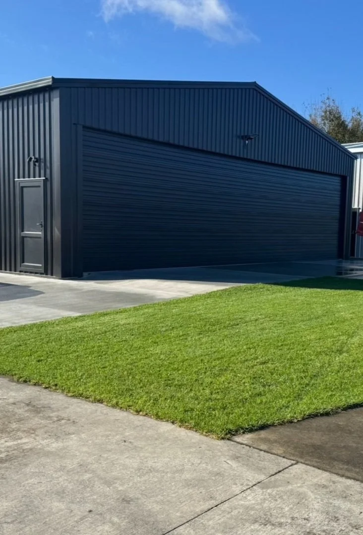 A black metal garage with a closed roll-up door, a side door, and a small security light, next to a patch of green grass and concrete pavement, under a partly cloudy blue sky.