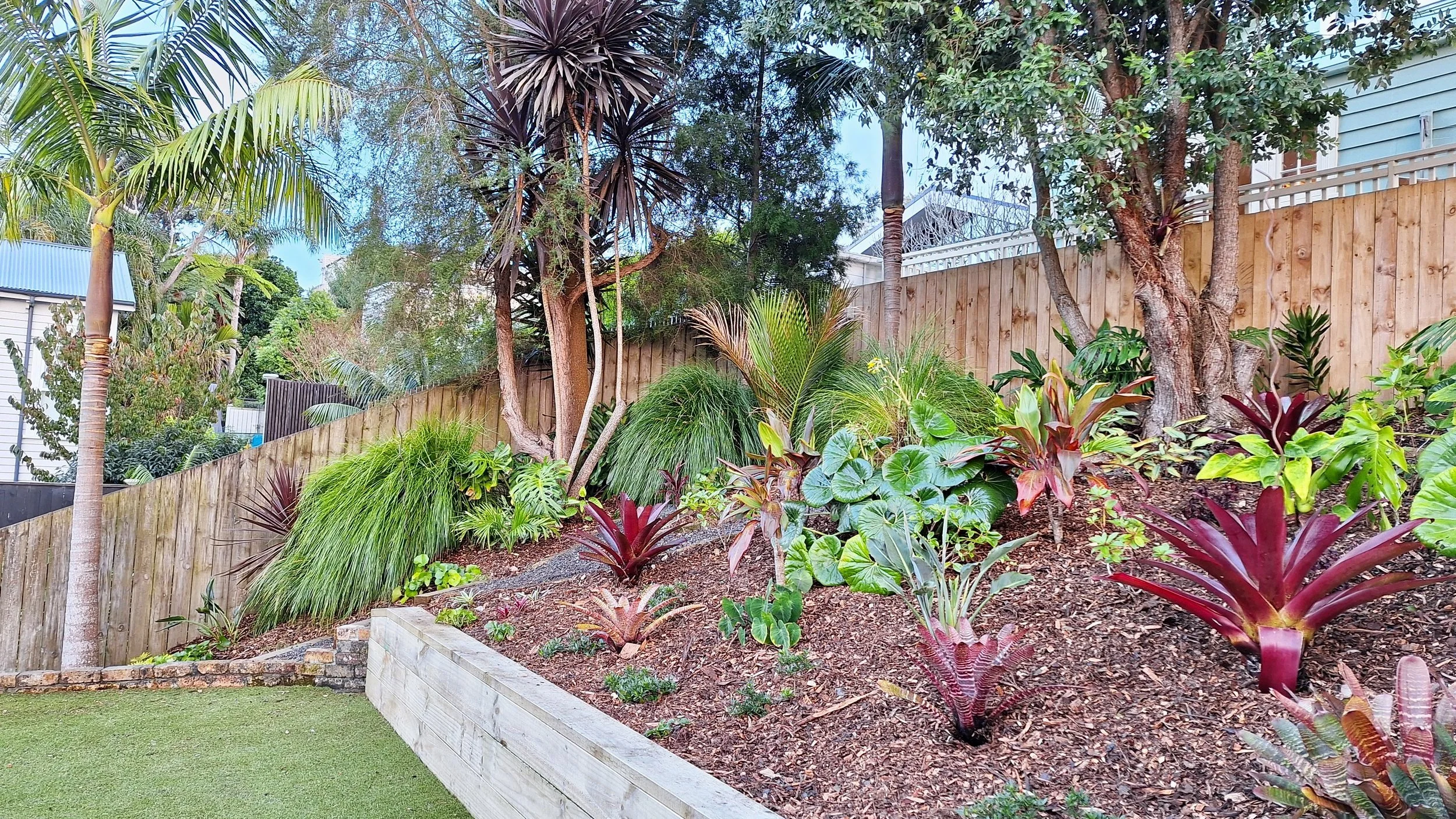 A landscaped backyard garden with various tropical plants, including palm trees, potted plants, and a wooden fence background.