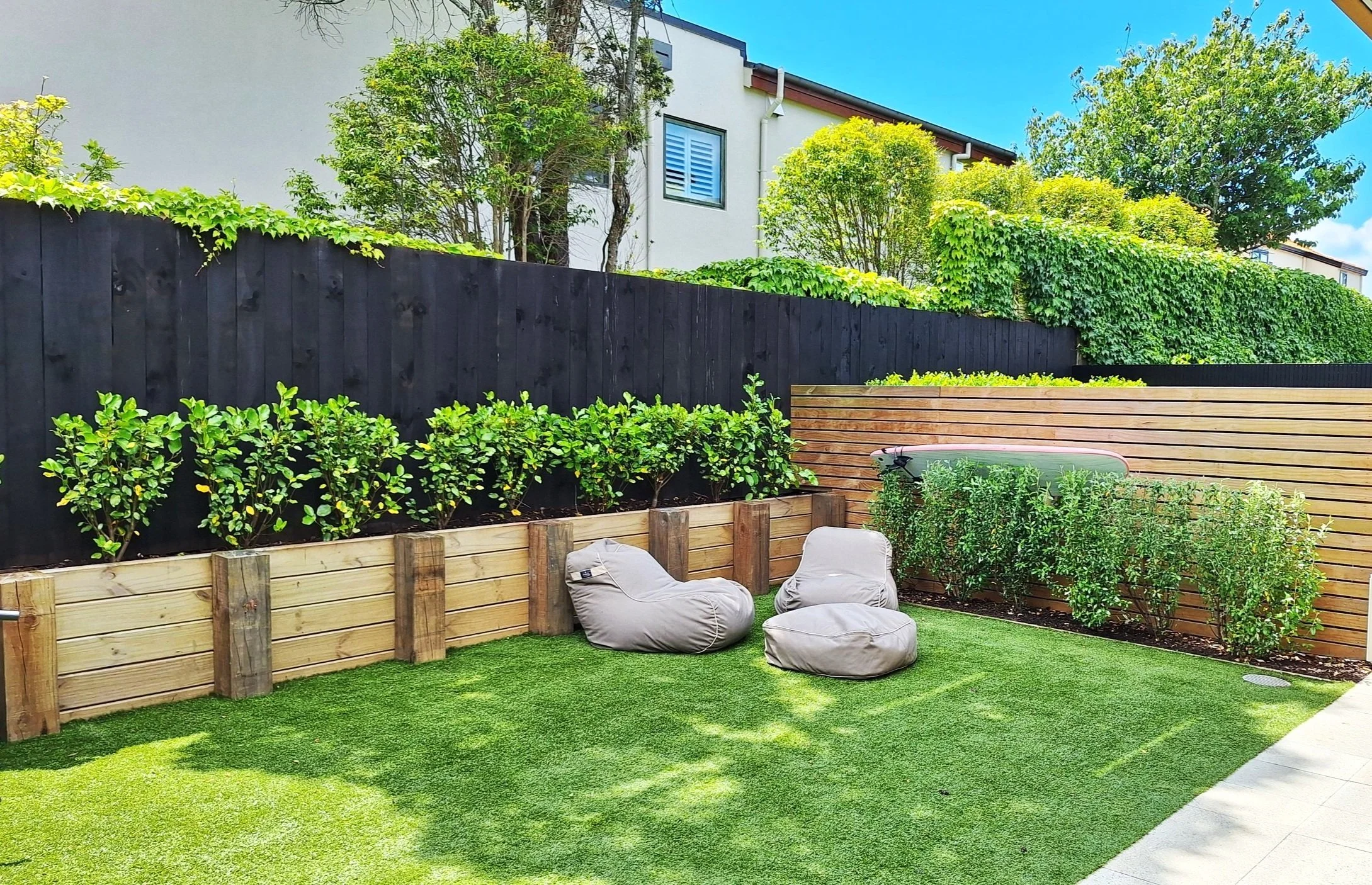 A backyard garden with green artificial turf, bean bag chairs, a hedgerow behind a wooden planter box, a fence with mature trees and multi-story buildings in the background, and a clear blue sky.