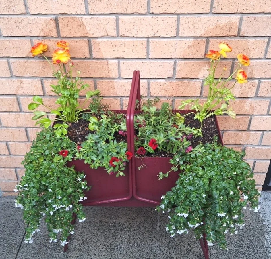 A metal flower stand with one containers filled with colorful blooming flowers, positioned against a brick wall outside.