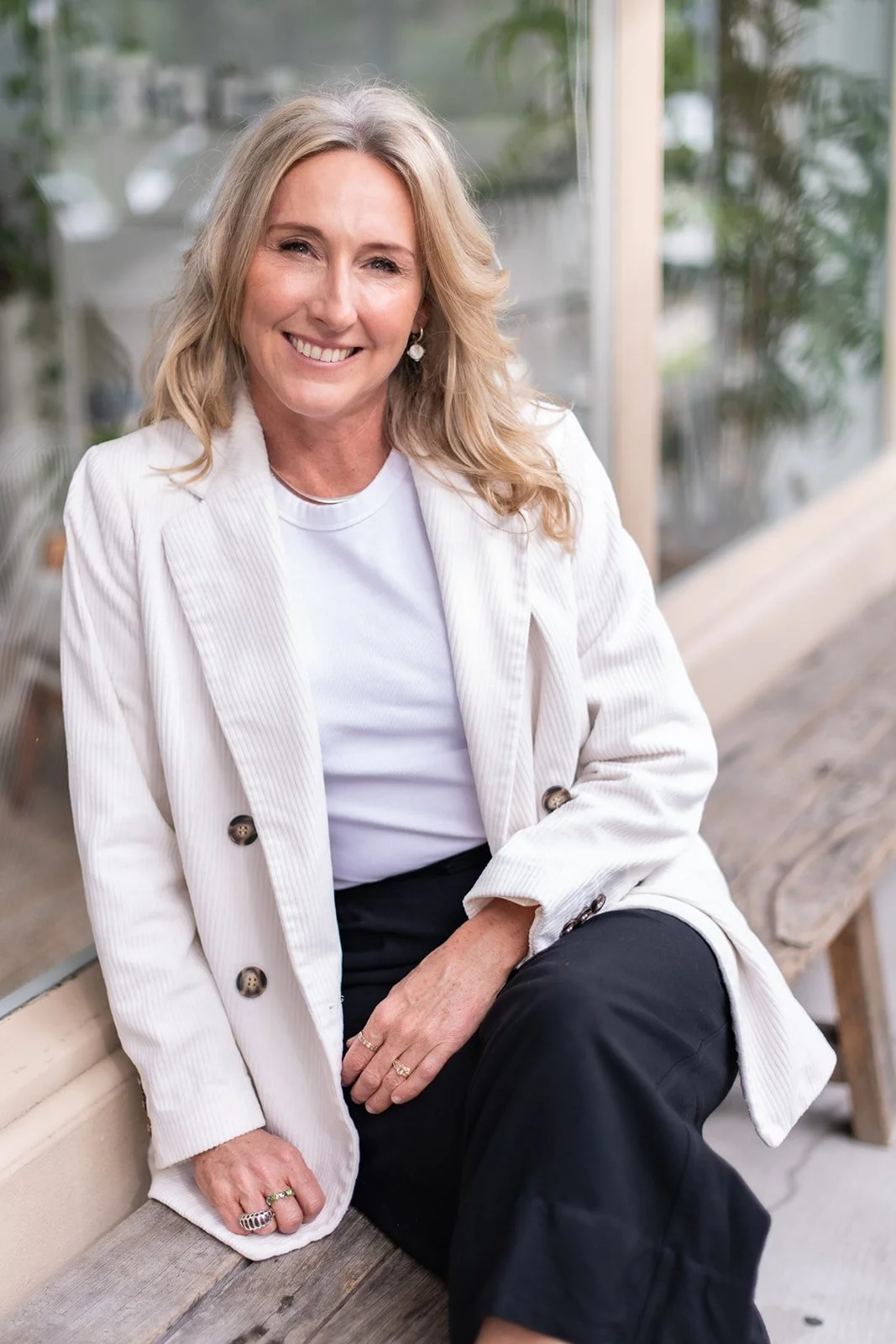 A smiling Naturopath specialising in iron imbalance disorder including anaemia and hemochromatosis is sitting on wooden bench with greenery in the background.