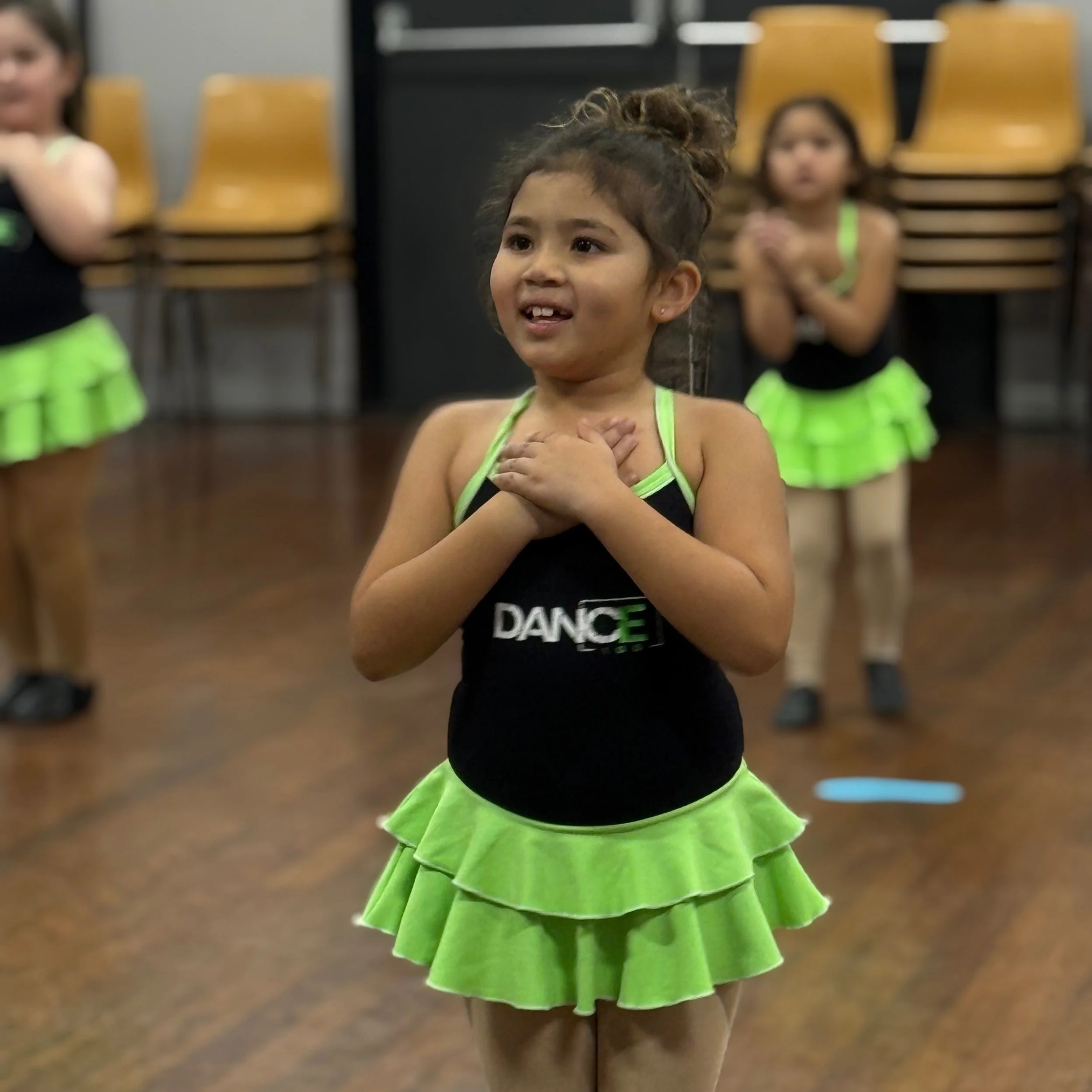 Young girl in a neon green dance costume with layered skirt, standing in a dance studio with her hands on her chest, smiling. Other children in similar costumes are in the background.