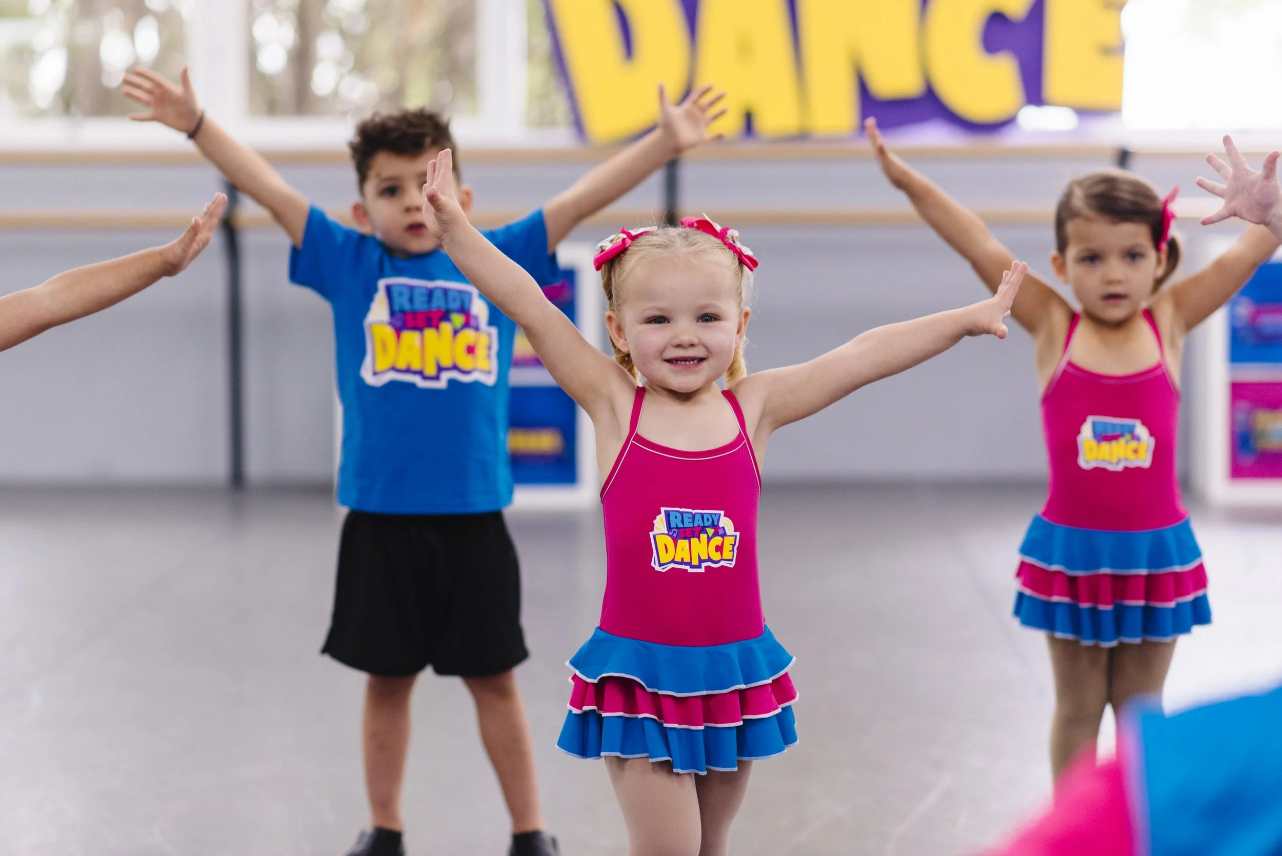Young children participating in a dance class, with arms raised and wearing colorful costumes. The background features a sign that says 'DANCE'.