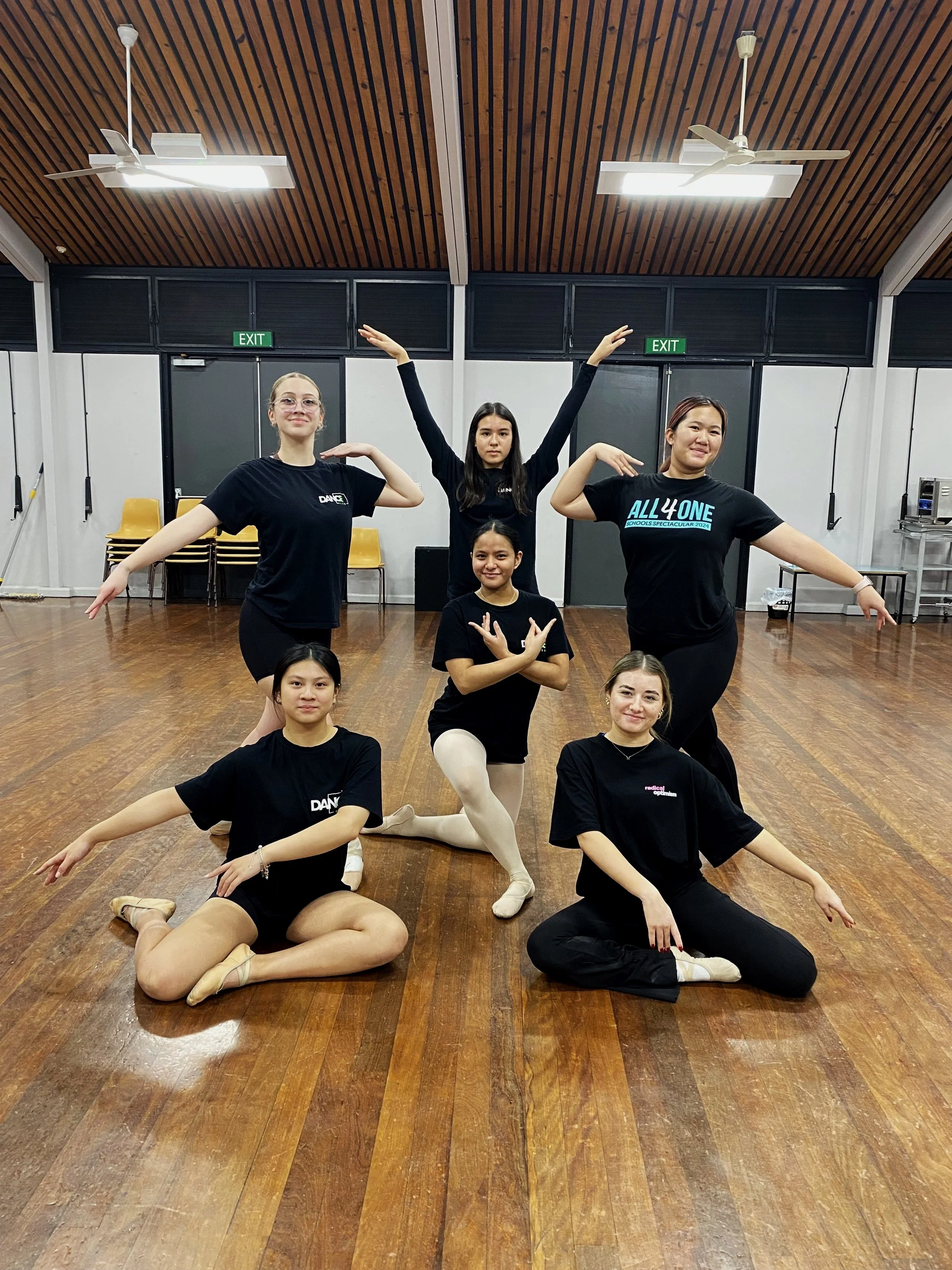 Six young women practicing dance or aerobics in a studio with wooden flooring, black walls, and a wooden ceiling, striking various poses.