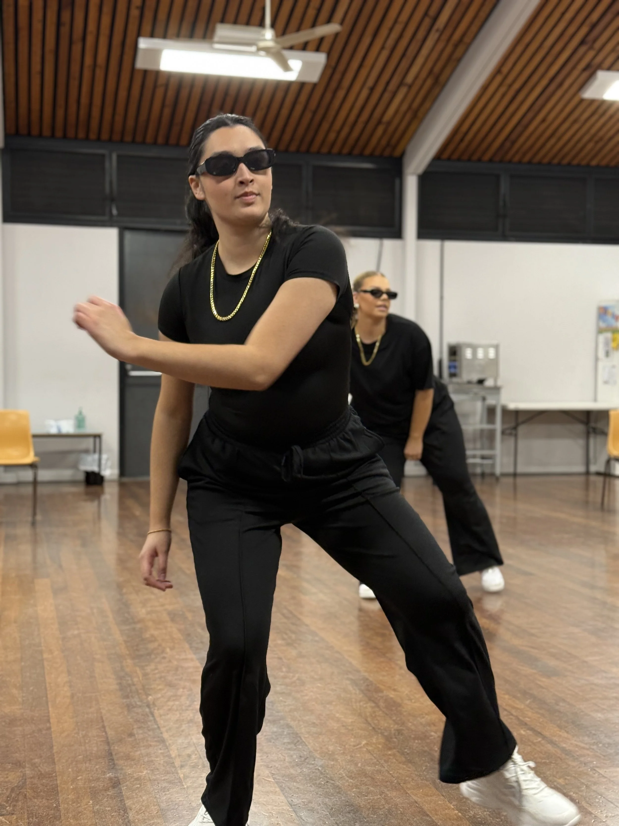 Two women dancing indoors, wearing black outfits, sunglasses, and gold chains, with a wood-paneled ceiling and wooden floor.