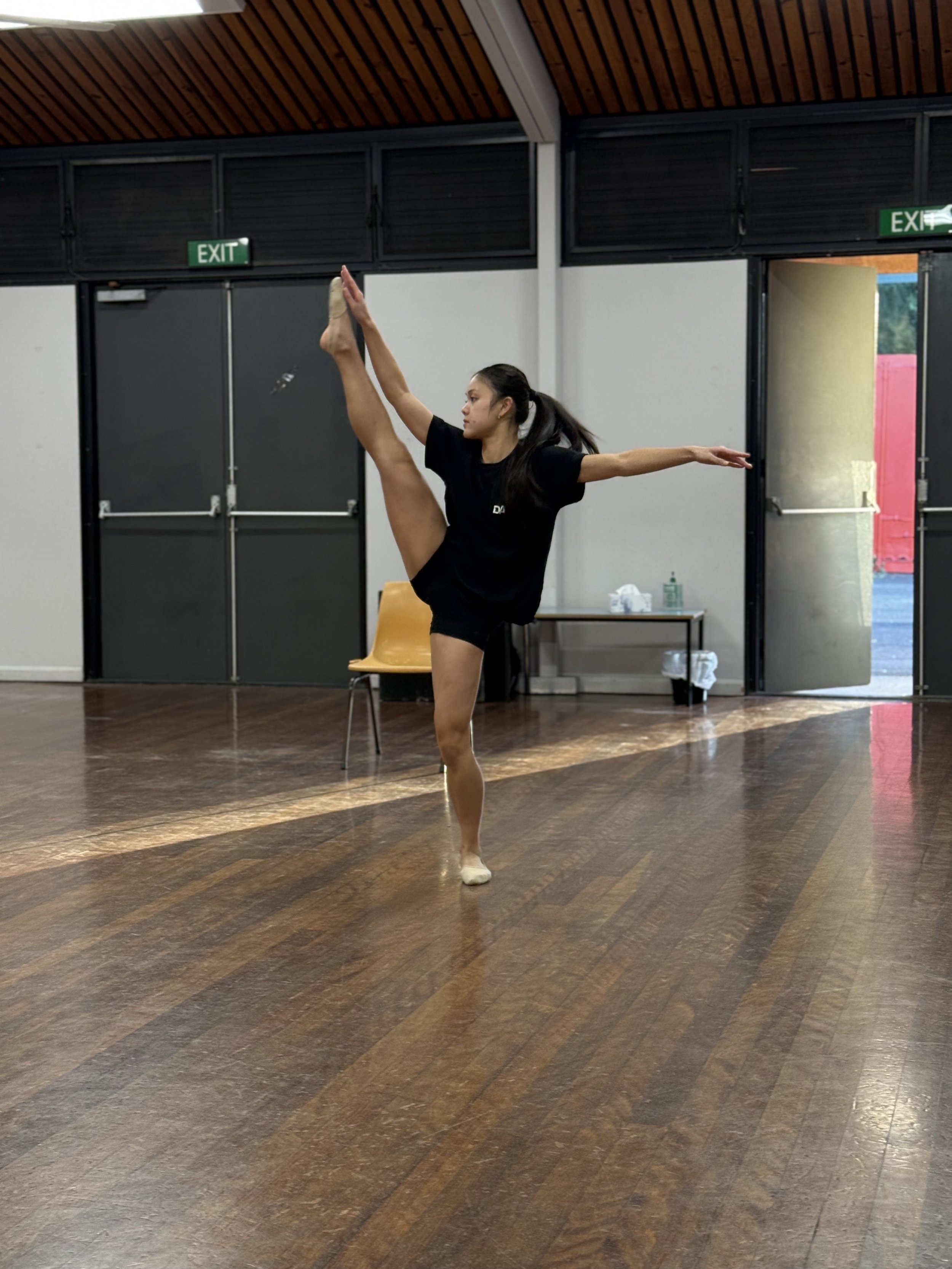 A woman in black athletic attire practicing ballet or dance in a studio, standing on one leg with the other leg extended high in front of her, with arms outstretched for balance.