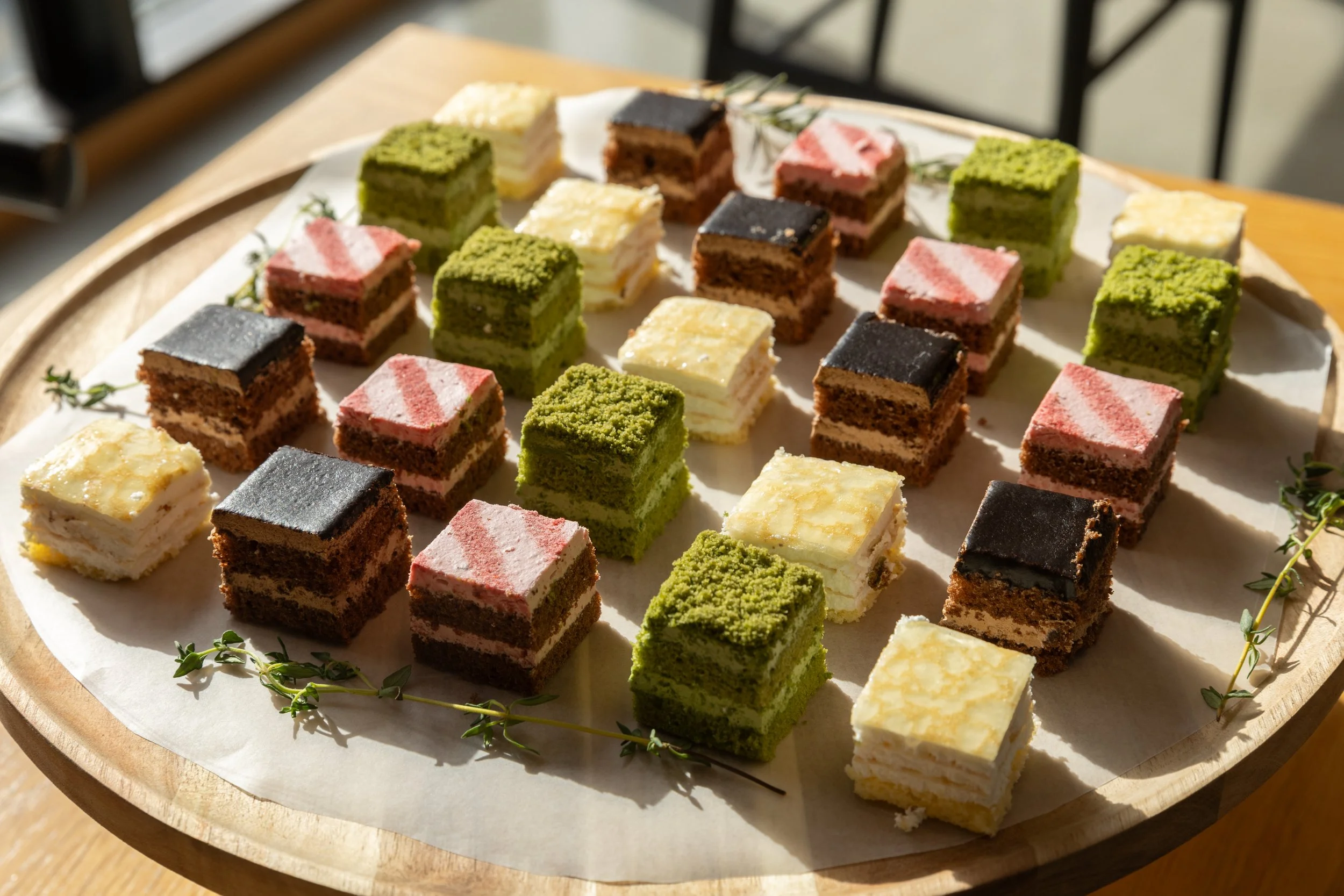 多層の小さなケーキが円形の木製トレイに並べられている。色とりどりのケーキは緑、ピンク、茶色、白色で、それぞれ層が見える。ケーキの間にハーブの枝が飾られている。