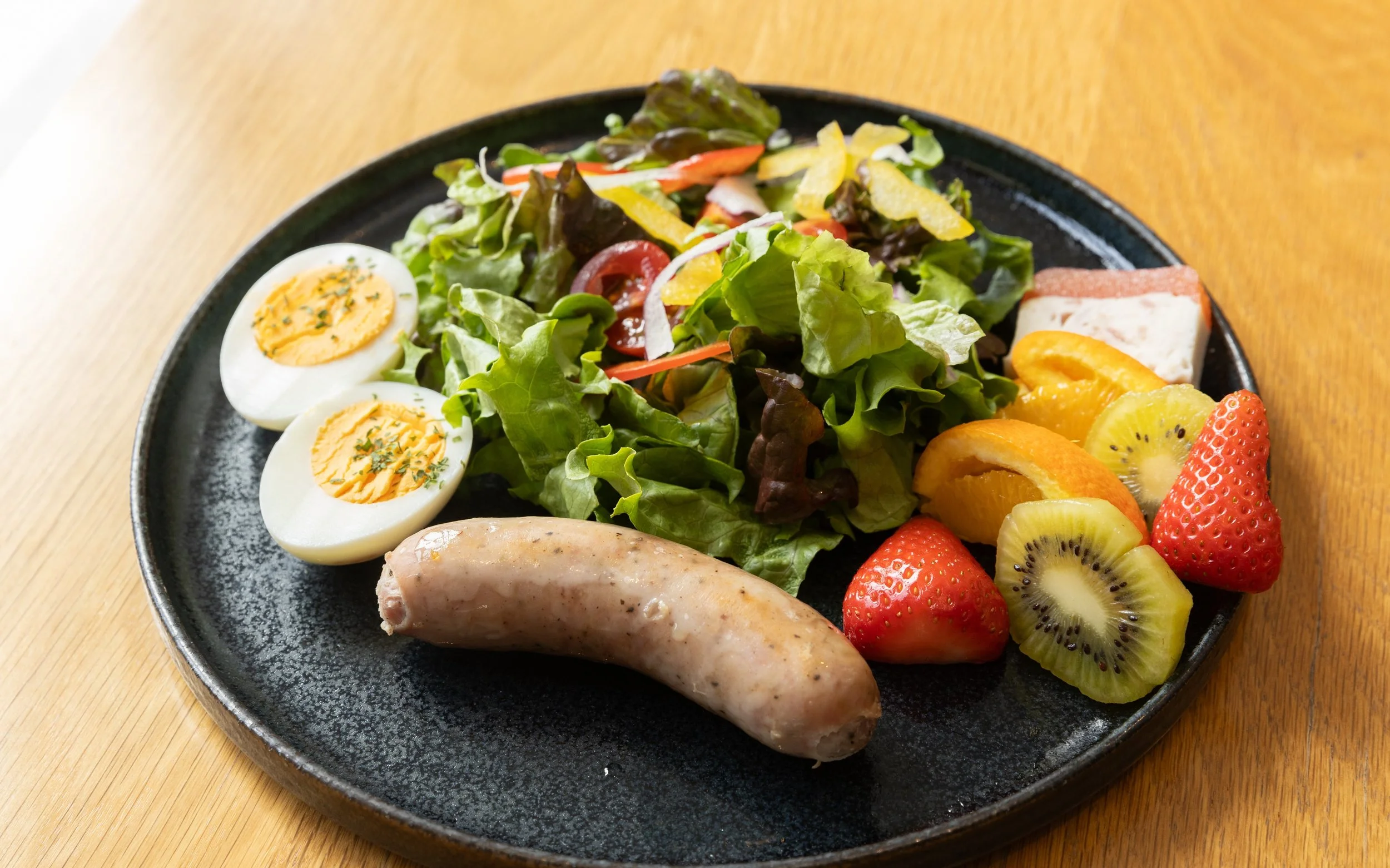 サラダ、ゆで卵、ソーセージ、フルーツの盛り合わせが黒いプレートにのっている