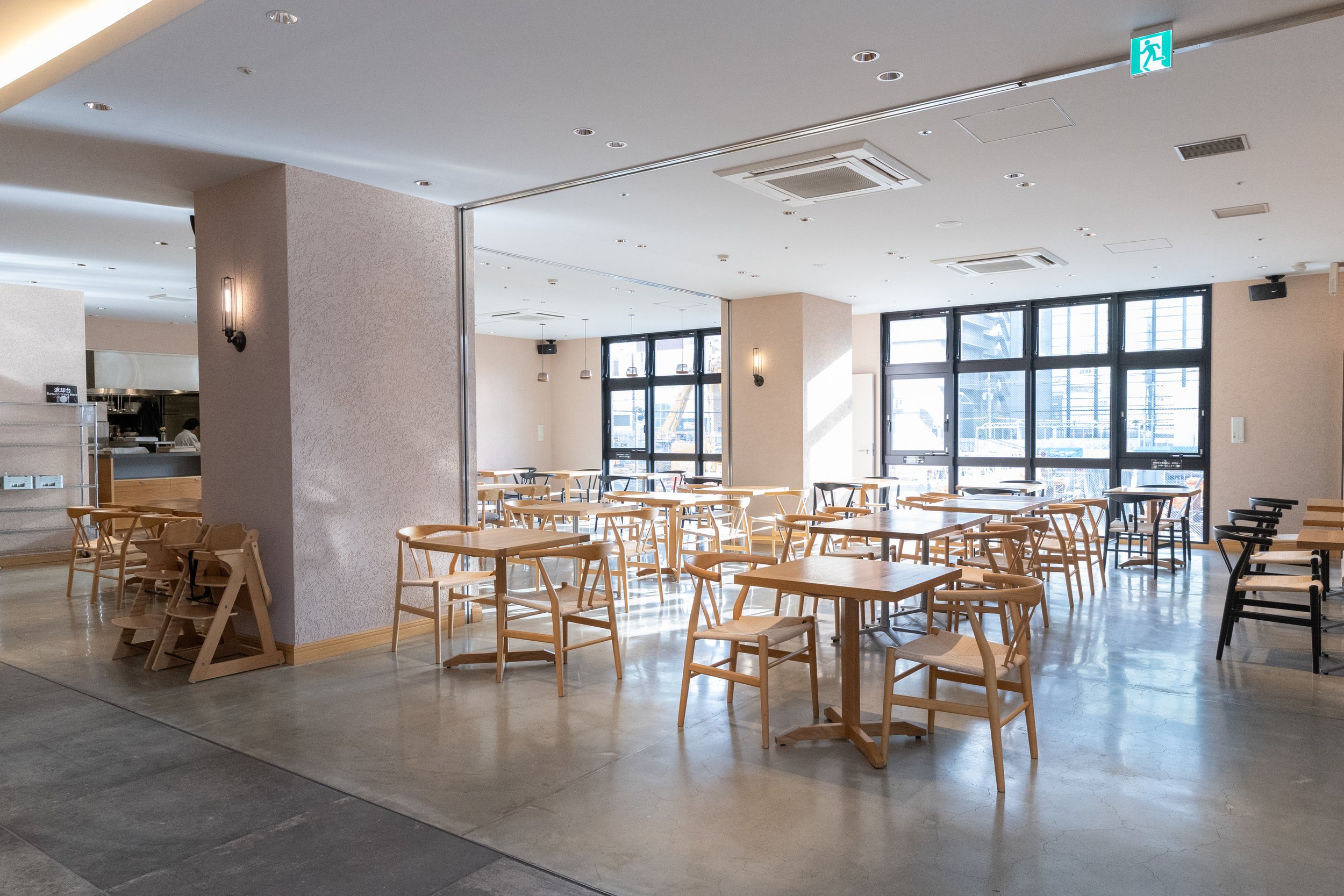 明るい自然光が入るレストランやカフェの空間。木製のテーブルと椅子が整然と配置されており、背後には大きな窓から外の景色が見える。
