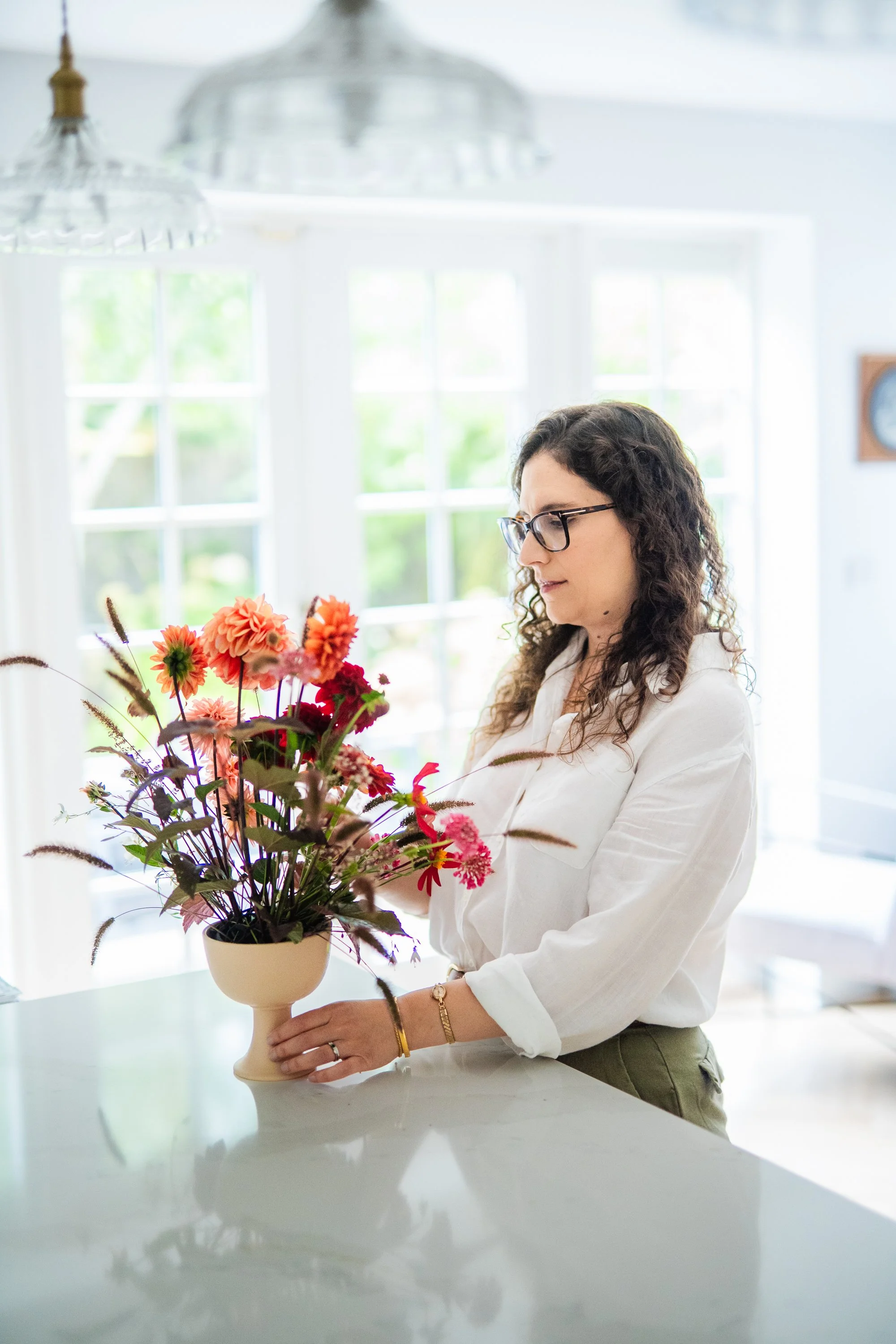 A woman with curly hair and glasses arranging a pink and orange flower bouquet in a beige vase on a white countertop in a bright room with large windows.