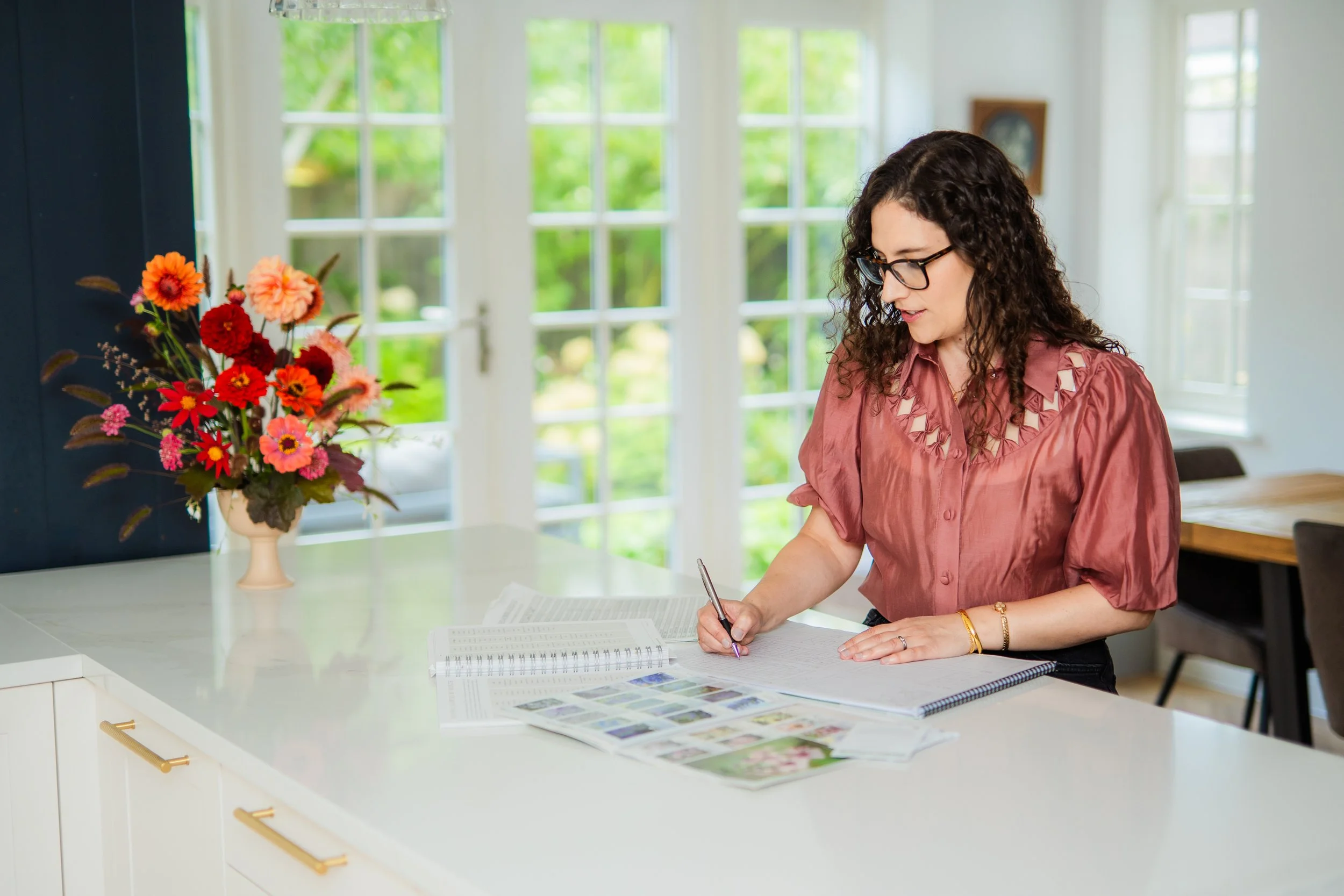 Woman with curly hair and glasses writing on paper at a kitchen island with a vase of colourful flowers, bright natural light coming through large windows.