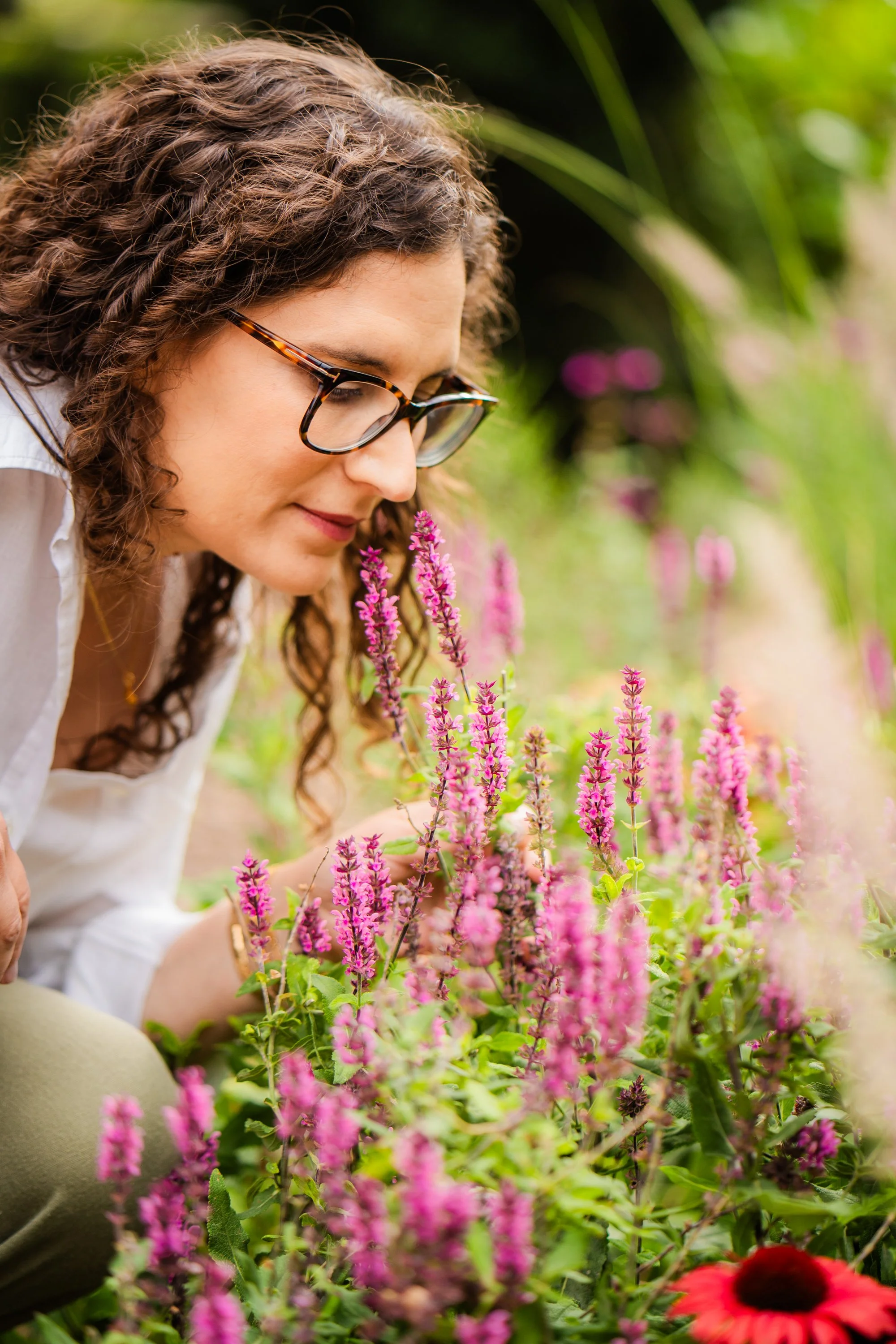 A woman with curly brown hair and glasses smelling pink salvias in a garden.