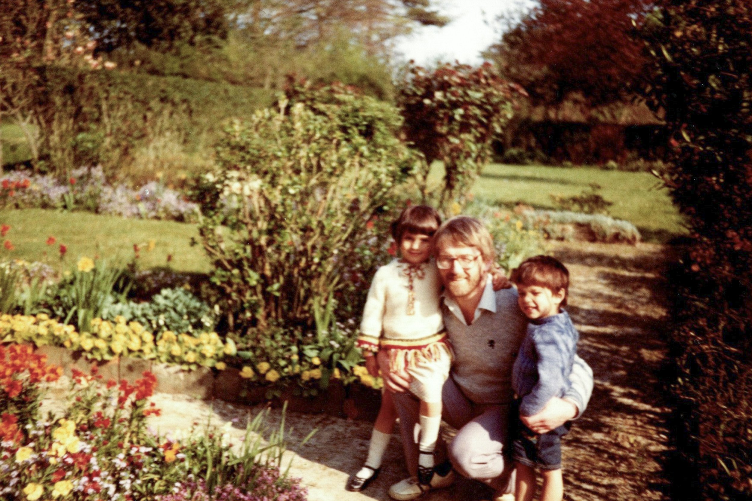 A man with two children in a garden with colorful flowers and bushes.